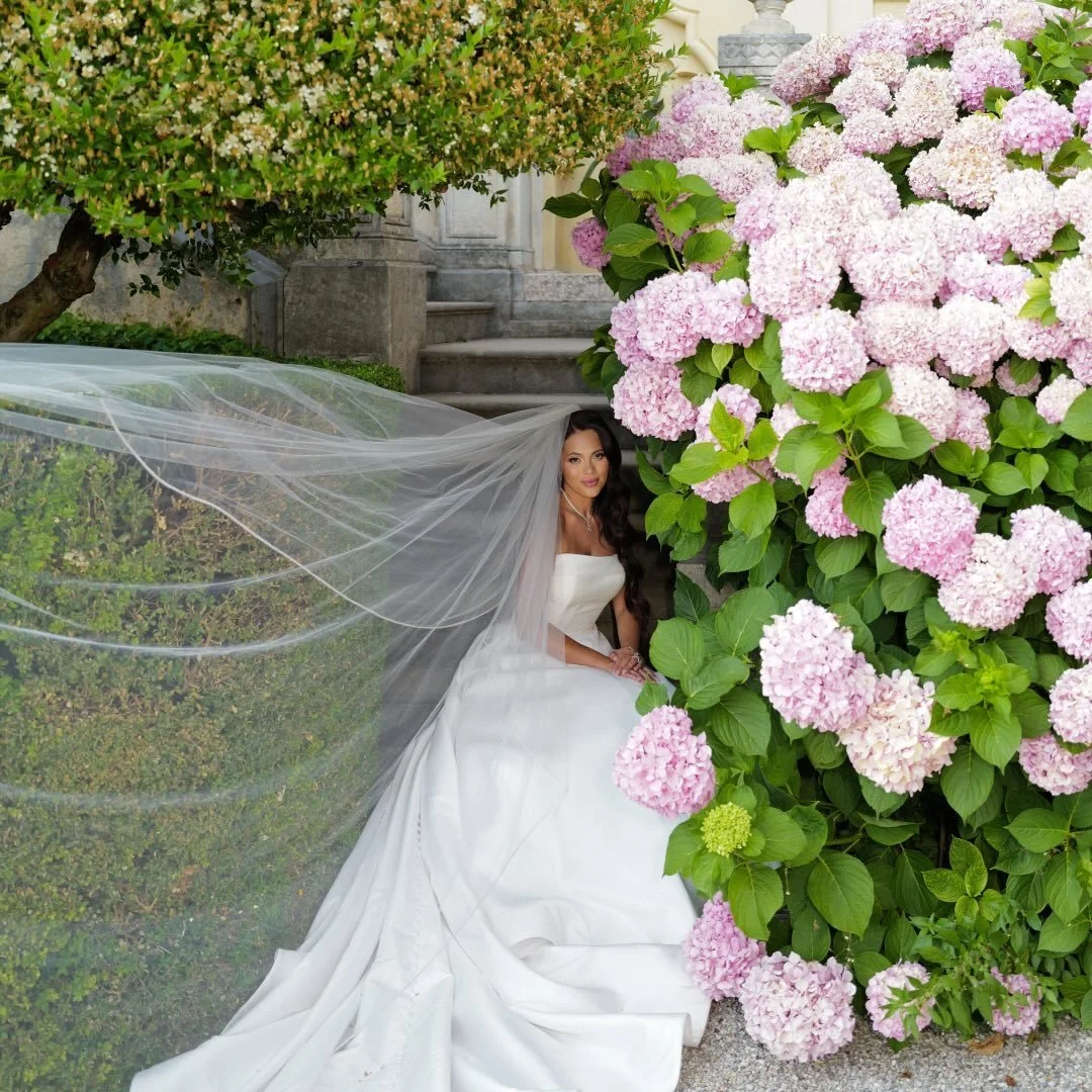 Bride in a white wedding gown and veil sitting beside pink hydrangea bushes and greenery.