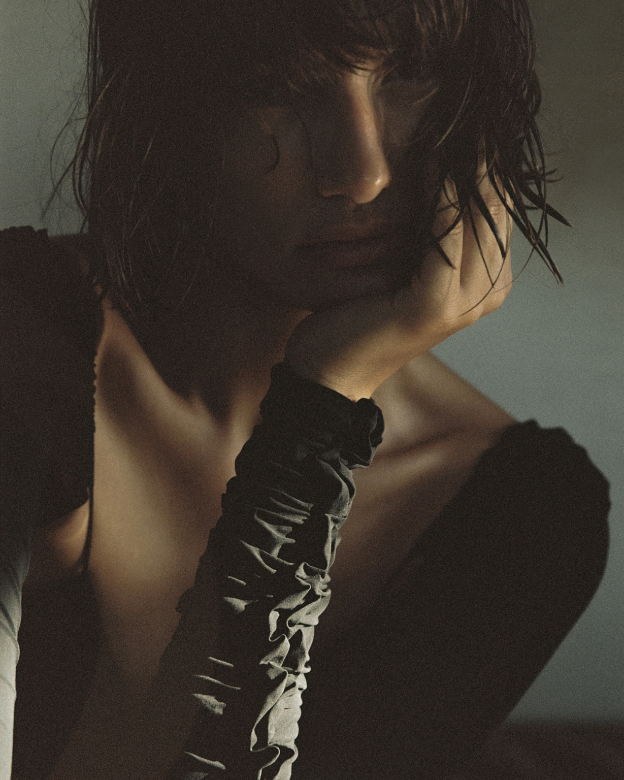 A woman with short, wet hair resting her head on her hand, wearing a black top and a scrunchie on her wrist, in dim lighting.