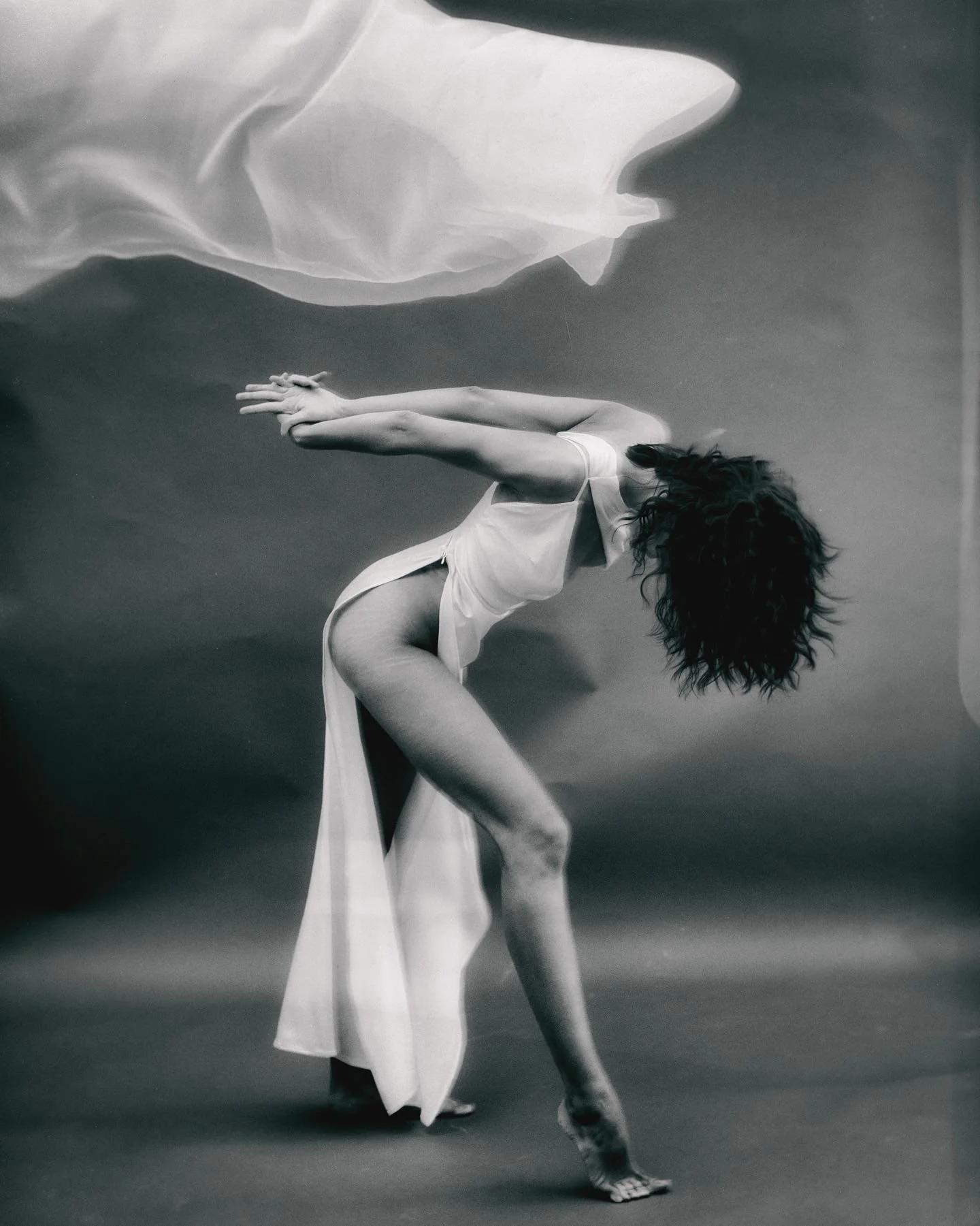 A black and white photograph of a female dancer in a bent-over pose, with her hands clasped behind her back, wearing a flowing dress with a slit, with a soft fabric above her head.