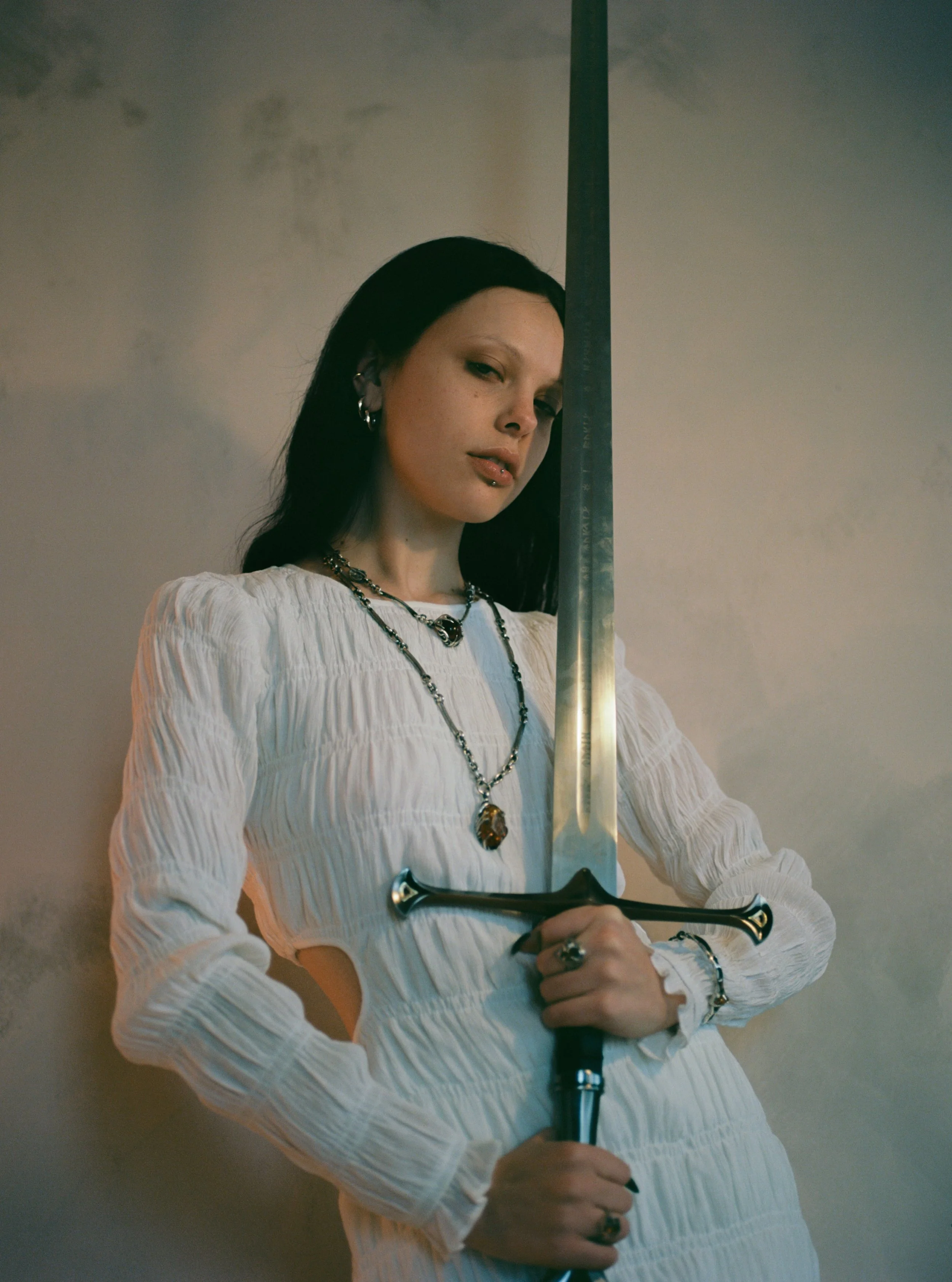 A woman with black hair is holding a large sword vertically in front of her, with part of the sword obscuring her face. She is wearing a white textured long-sleeve blouse and various jewelry including necklaces and rings, against a neutral background.