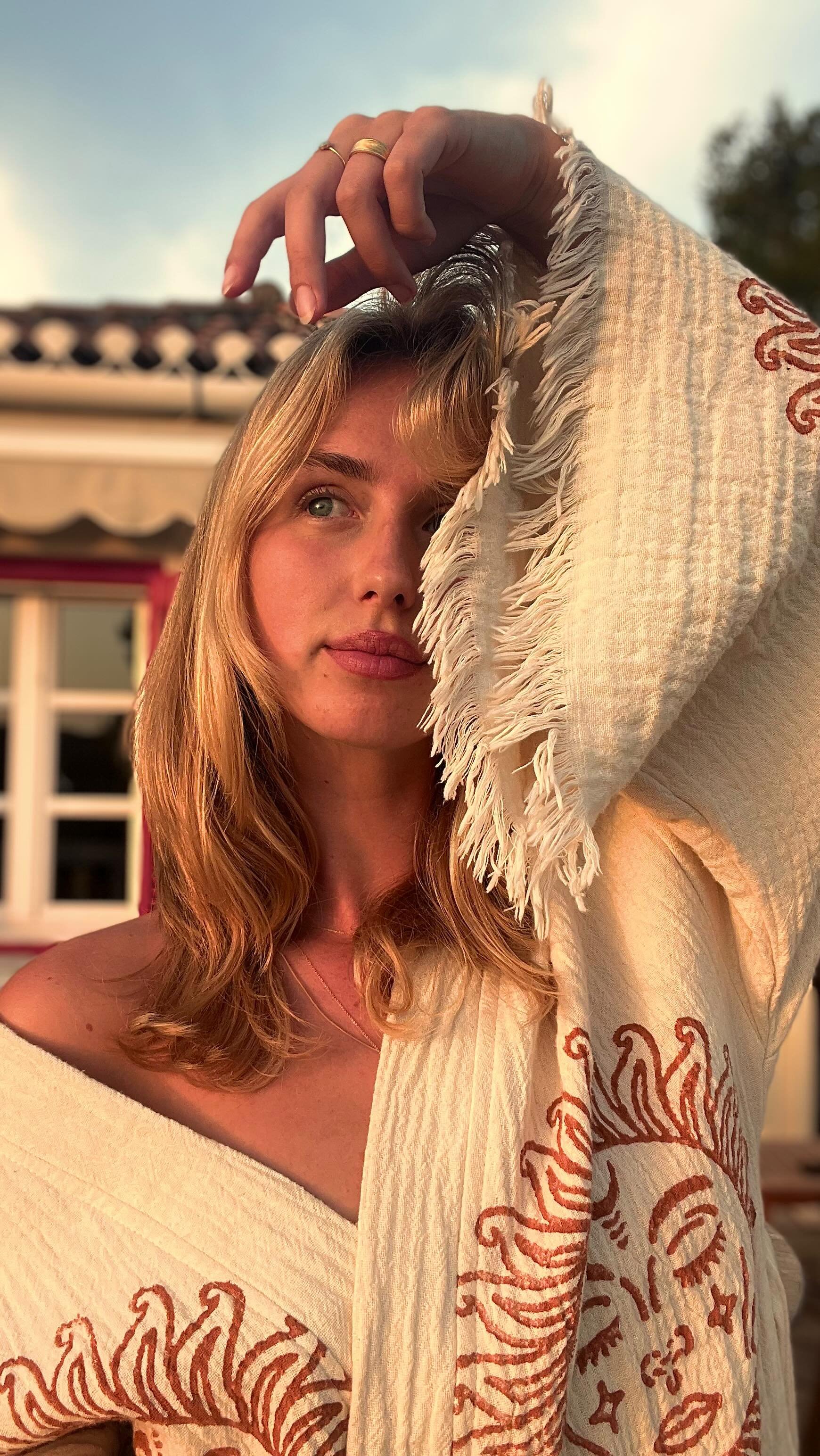 A young woman with wavy blonde hair and blue eyes is holding a fringed towel over her head, looking into the distance during sunset. She wears a beige off-shoulder top with red decorative patterns.