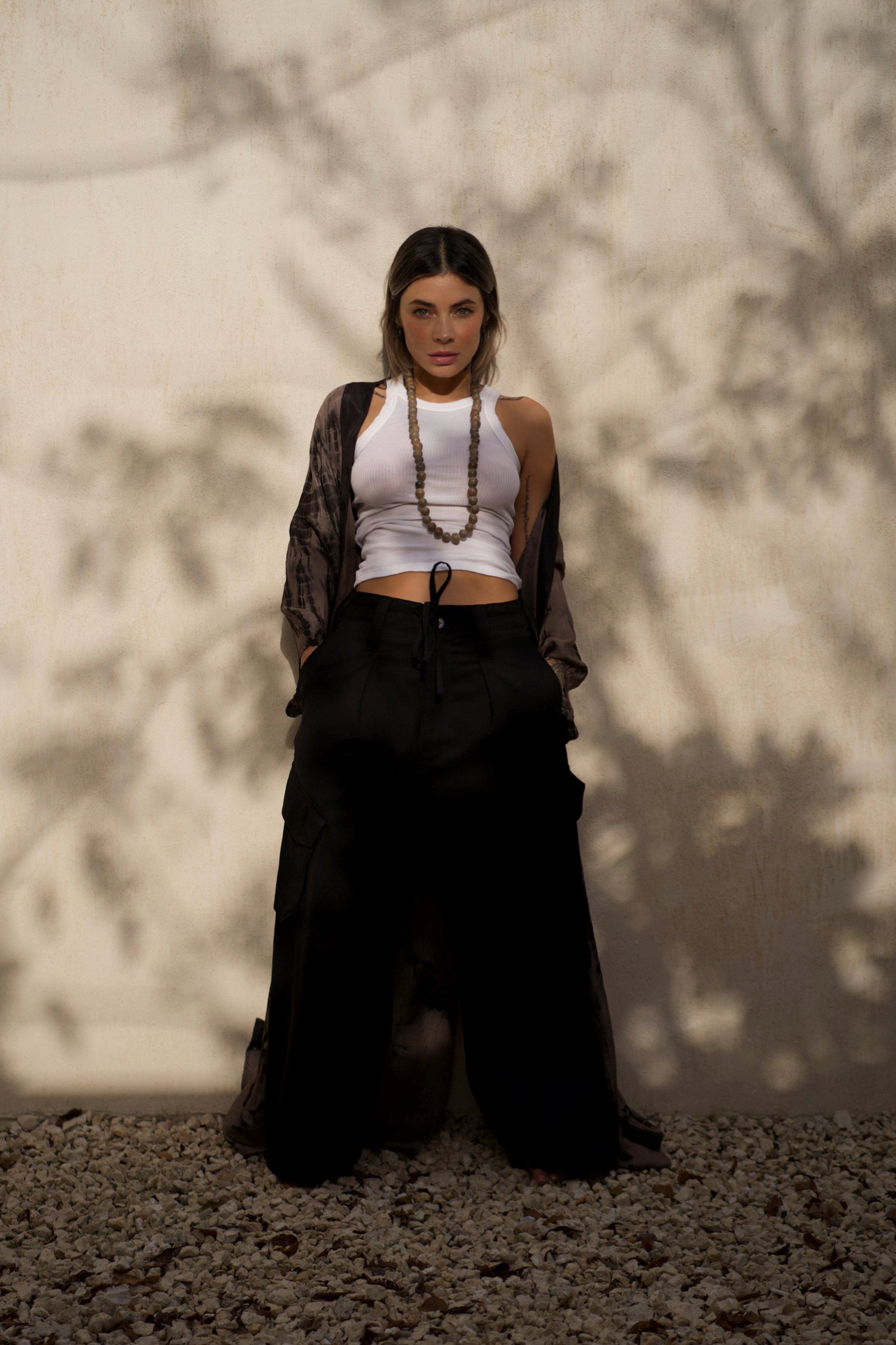 A young woman with shoulder-length dark hair and light makeup, standing against a textured beige wall, with shadows of leaves cast behind her. She's wearing a white sleeveless crop top, black loose-fitting cargo pants, and a long beaded necklace. She has a dark jacket draped over her shoulders and stands with her hands in her pockets.