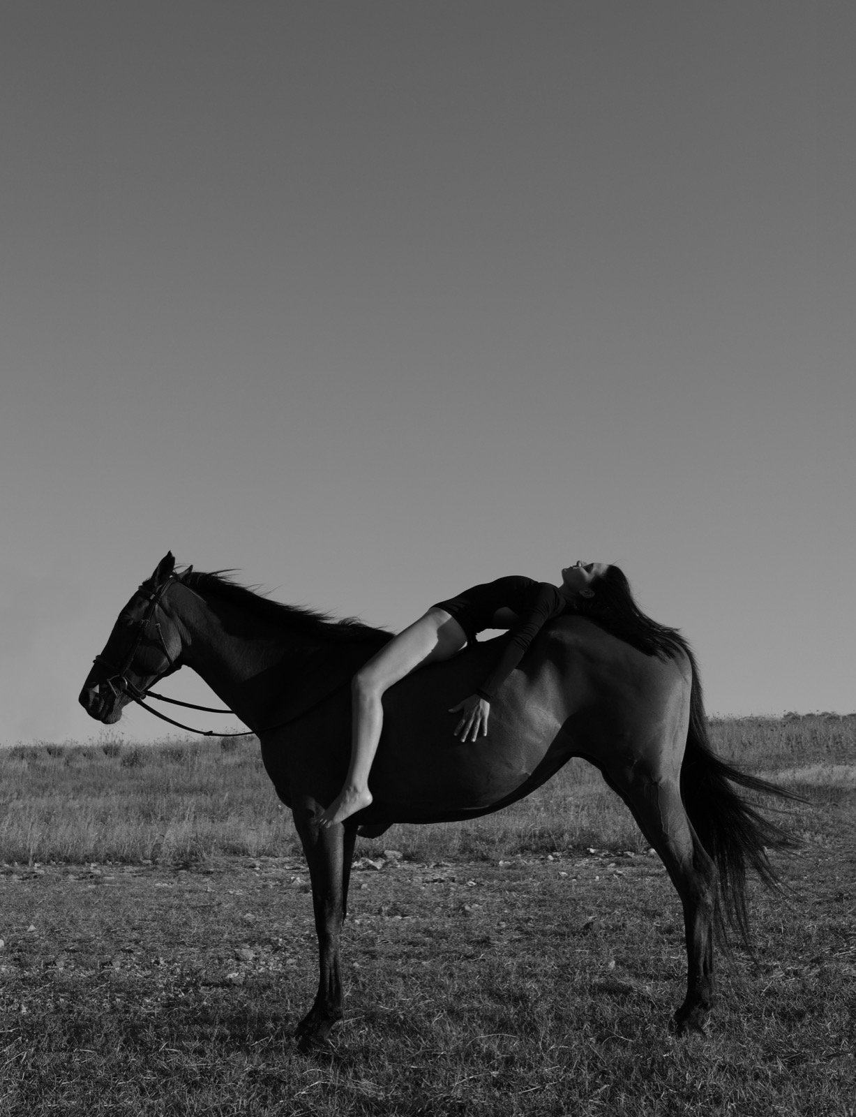 A woman in ballet attire lying backward on a rearing horse outdoors on a grassy plain, with a clear sky in the background.
