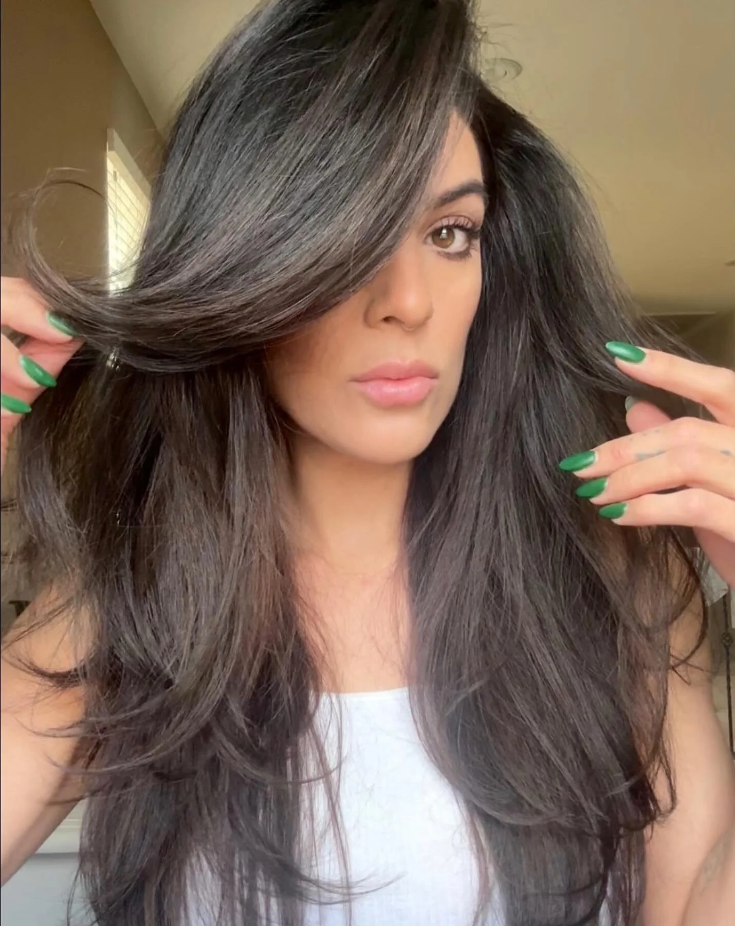 A woman with long, dark brown, wavy hair partially covering her face, holding hair with both hands, wearing a white top and green nail polish, in a room with a window and beige walls.