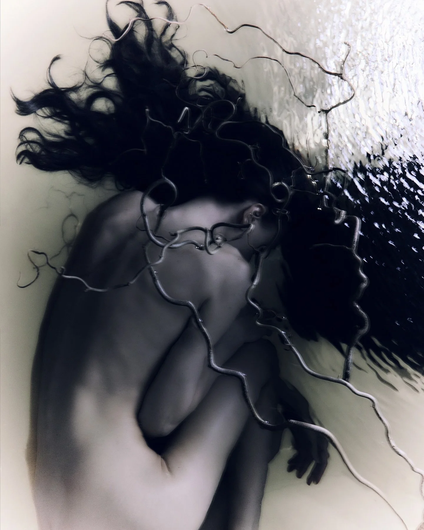 Black and white photo of a woman submerged in water, with her body curled and her face turned downward, surrounded by tangled, thin branches or wires.