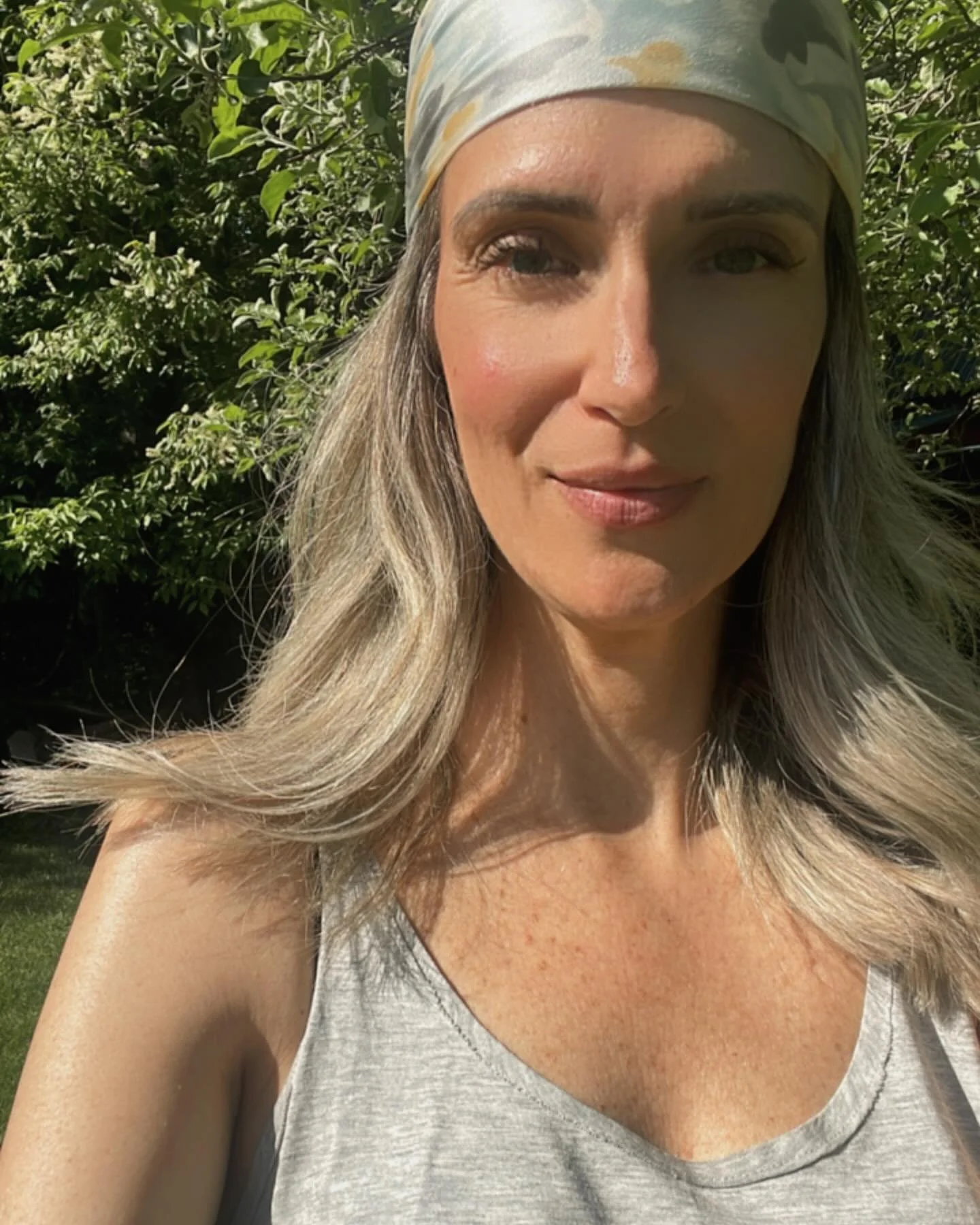 A woman with blonde hair wearing a patterned headscarf and gray tank top, taking a selfie outdoors with green foliage in the background on a sunny day.