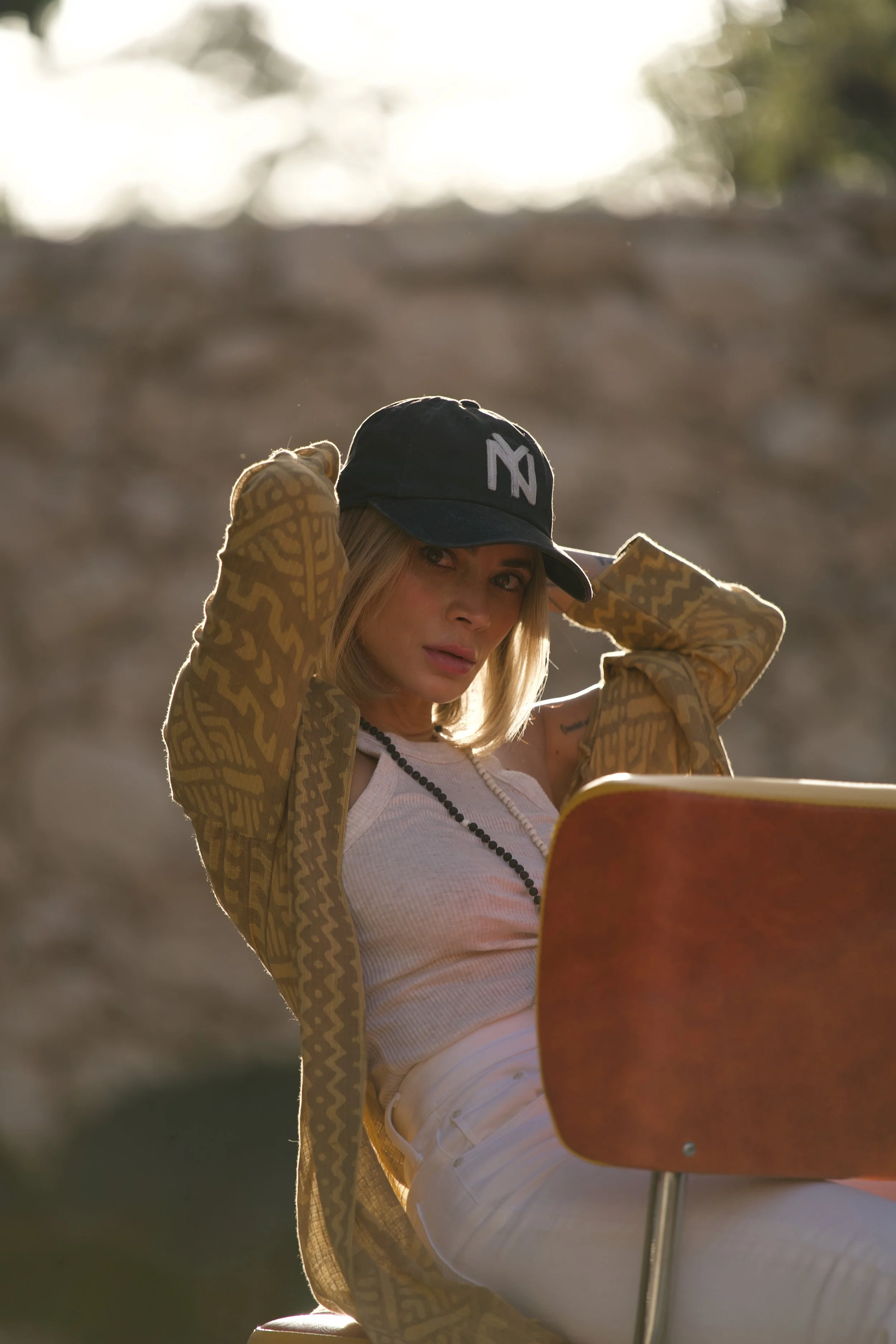 A woman wearing a black cap, a tank top, and a patterned jacket, sitting outdoors during the day, adjusting her cap, with sunlight in the background.