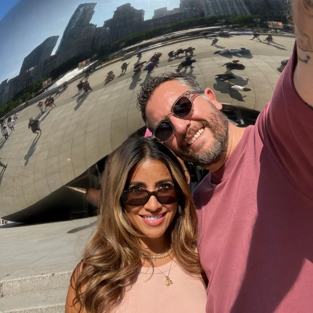 A smiling couple takes a selfie in front of Cloud Gate, also known as The Bean, in Millennium Park in Chicago, Illinois. The reflection in the sculpture shows city skyscrapers and visitors.