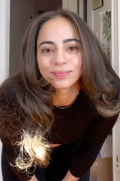 A young woman with long, wavy brown hair smiling slightly at the camera in a cozy indoor setting.