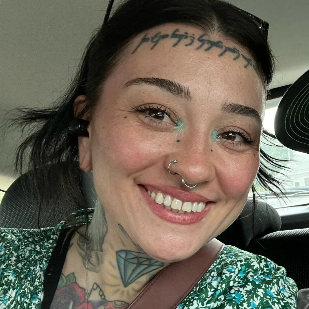 Close-up of a smiling woman with tattoos, piercings, and makeup, sitting in a car.