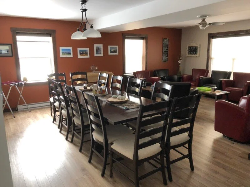 A dining and sitting area with a long table surrounded by chairs, and a separate seating area with multiple armchairs and sofas, with windows allowing natural light to fill the room.