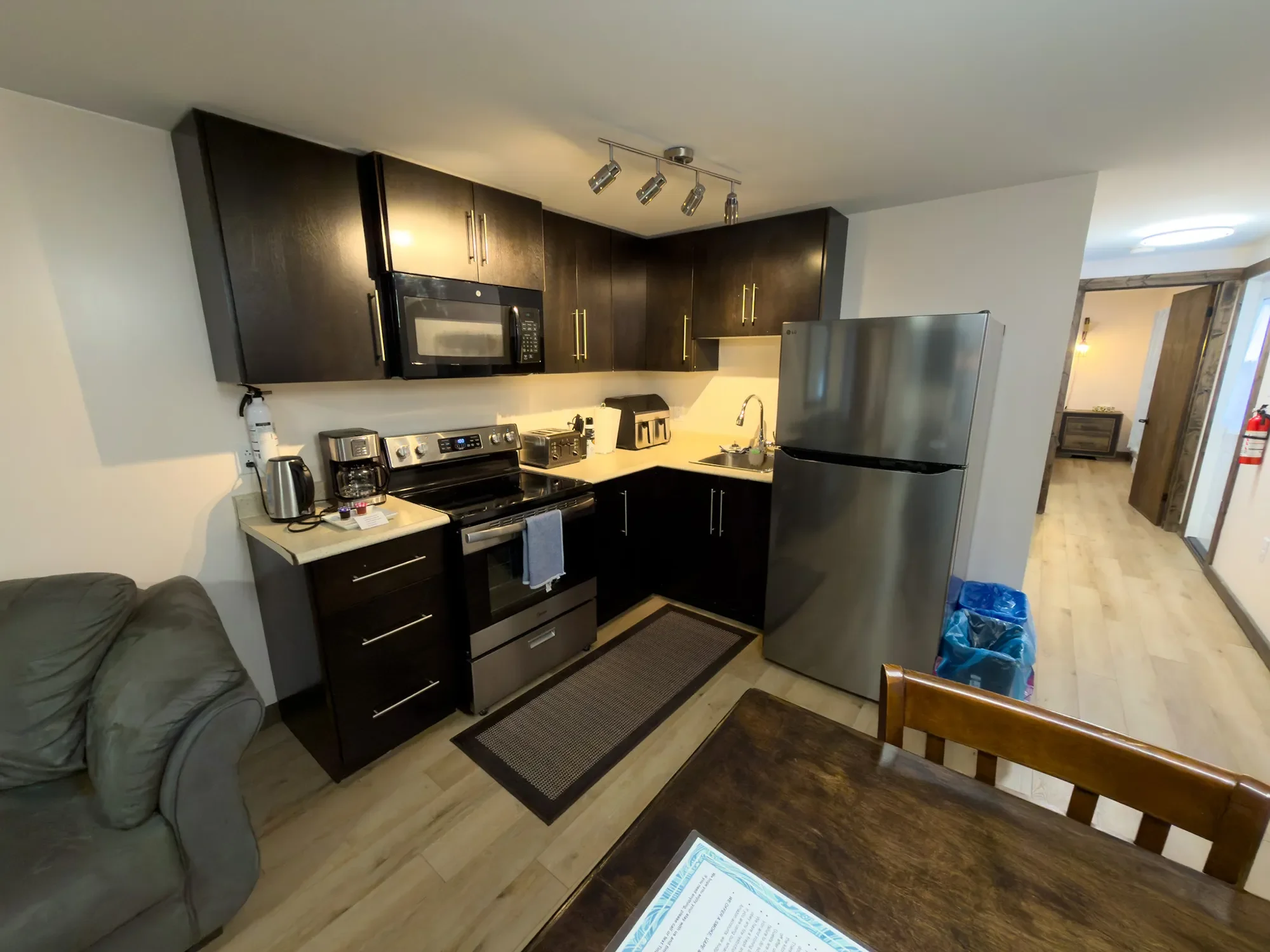 Kitchen with dark cabinets, stainless steel refrigerator, microwave, stove, and various appliances on the counter. A small dining table in the foreground and a hallway with wood flooring in the background.