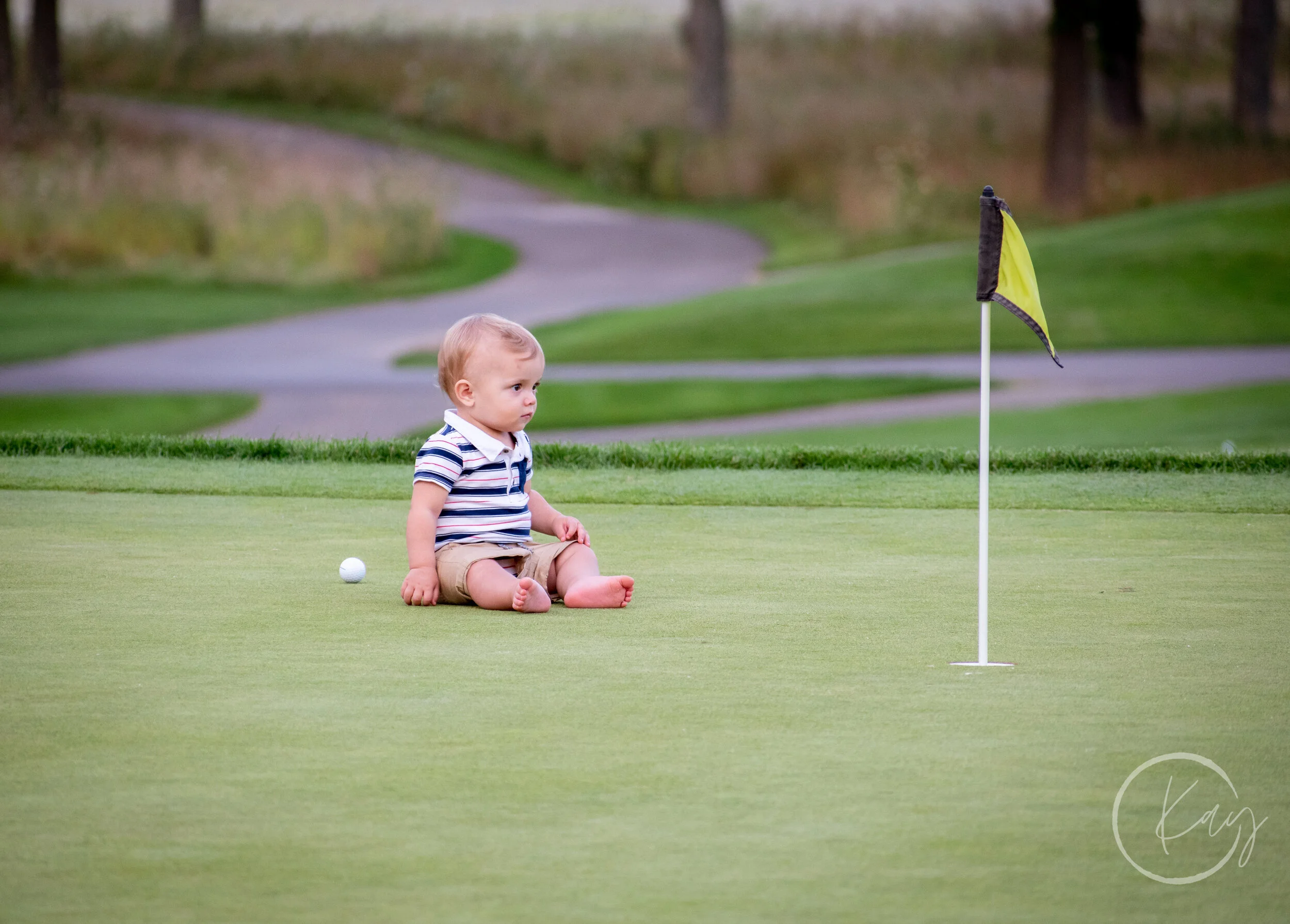 A young child sitting on a golf green near a golf hole with a flag, looking thoughtfully at the hole, with trees and a pathway in the background.
