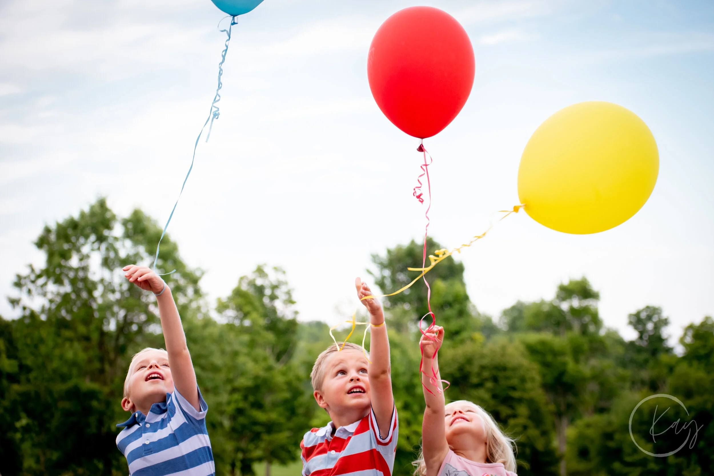 Three children playing outdoors, releasing red, yellow, and blue balloons into the sky during daytime.