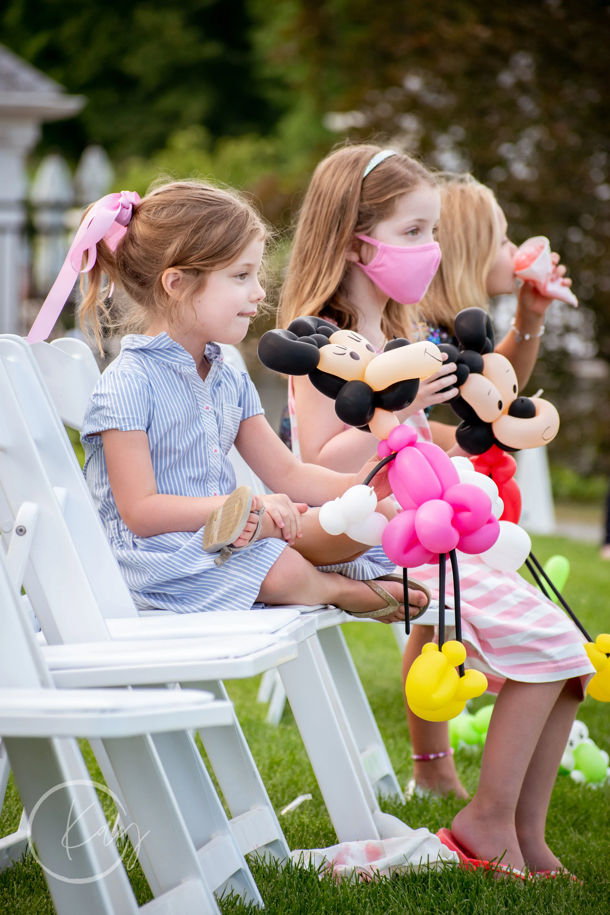 Three young girls seated on white folding chairs at an outdoor party, holding balloon characters of Mickey Mouse and Minnie Mouse, with one girl drinking from a bottle.