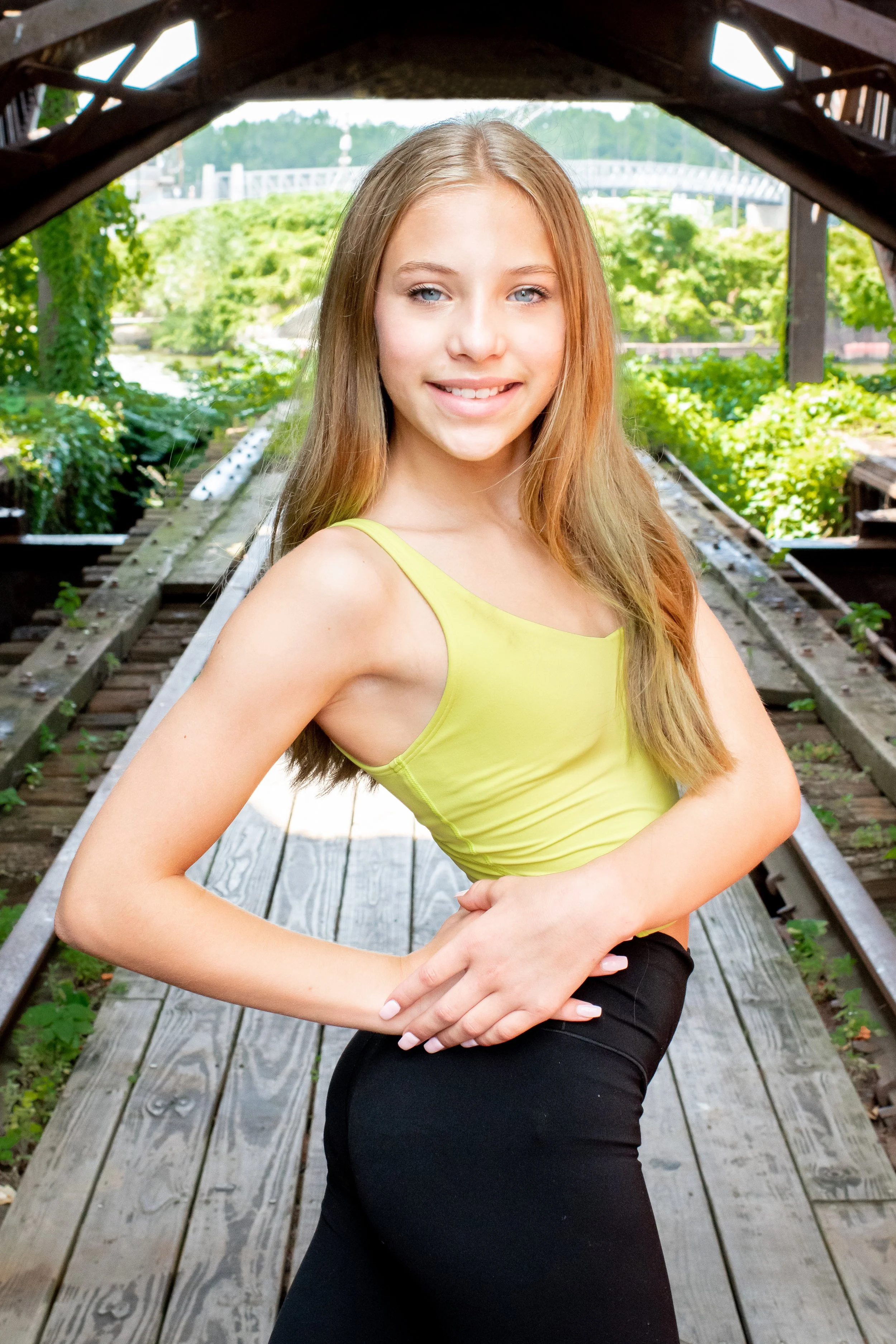 A young woman with long, light brown hair and blue eyes posing with her hands on her waist on a wooden bridge over a creek, surrounded by green trees and vegetation, with a bridge visible in the background.