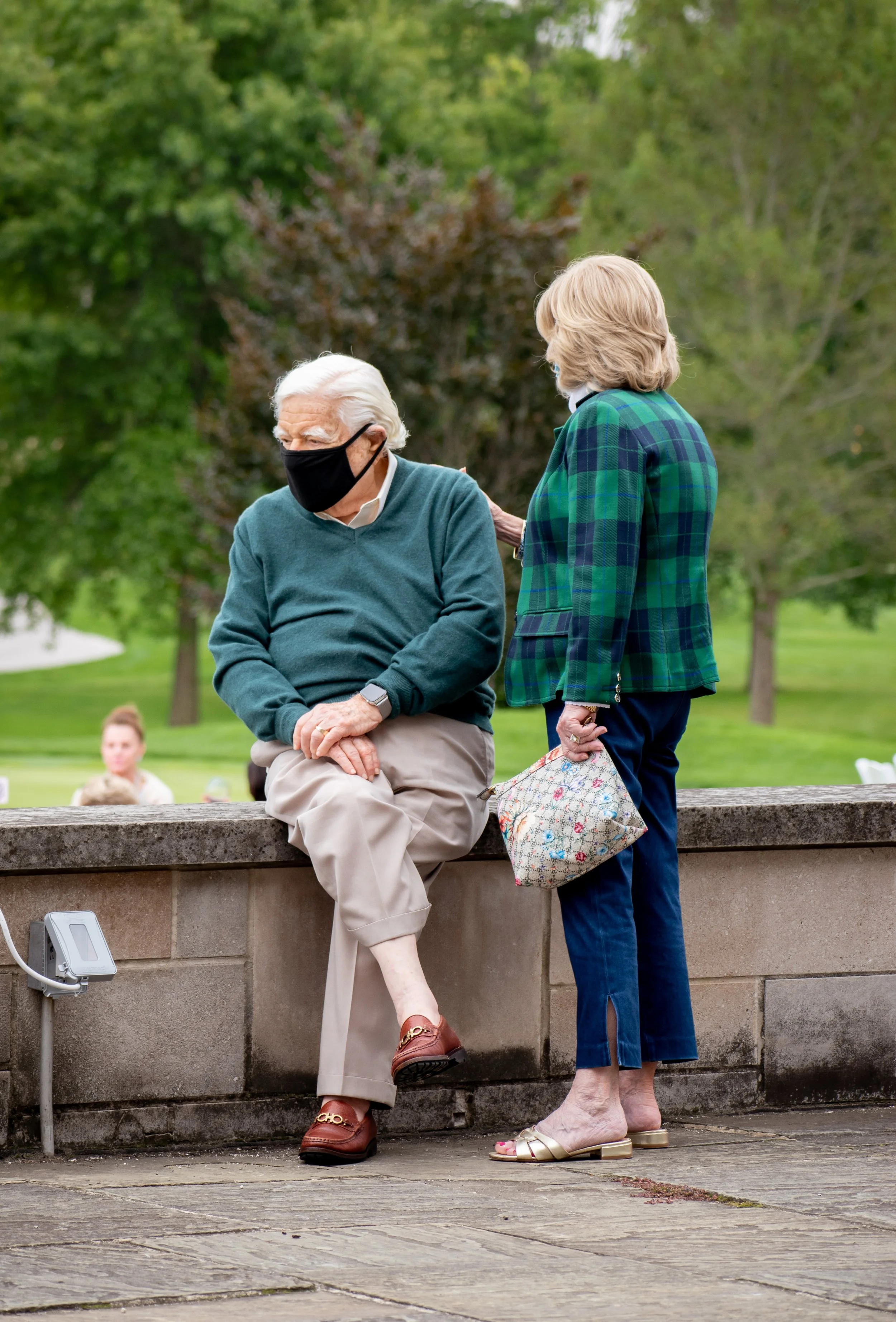 An elderly man with white hair and a face mask sits on a stone ledge, talking to a woman with blonde hair who stands nearby in a park, holding a patterned handbag and wearing open-toe sandals. The background includes trees and greenery.