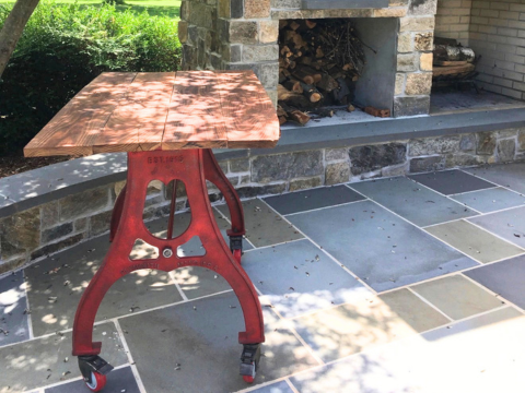 Red cast iron counter height table base with wood top on a stone patio next to an outdoor fireplace