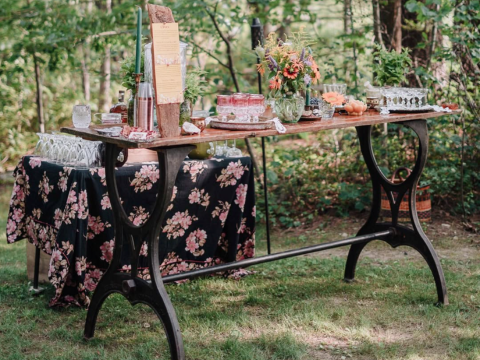 Black cast iron bar height table base outdoors with wood top, floral tablecloth, and flower arrangements