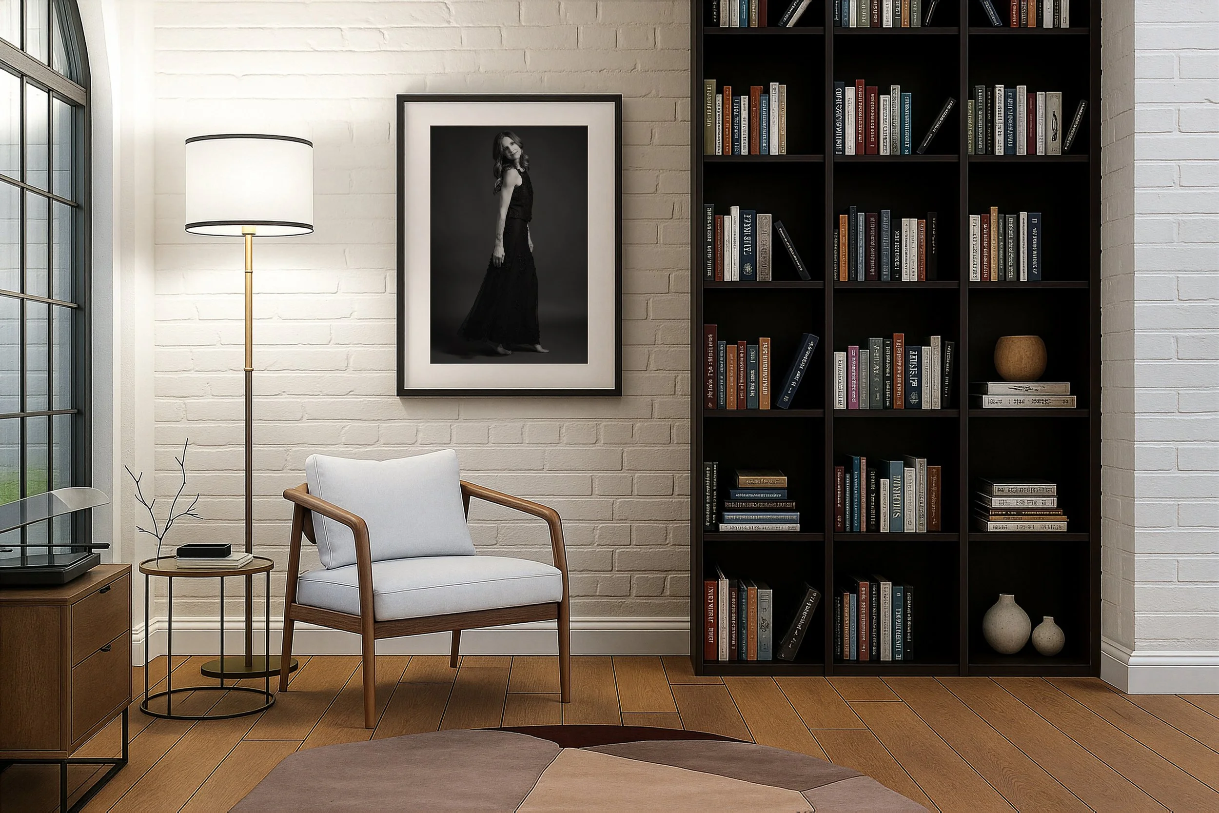 Modern living room with a white brick wall, bookshelf filled with books, black and white portrait photograph, white armchair with wooden arms, floor lamp, and part of a window with a grid pattern.