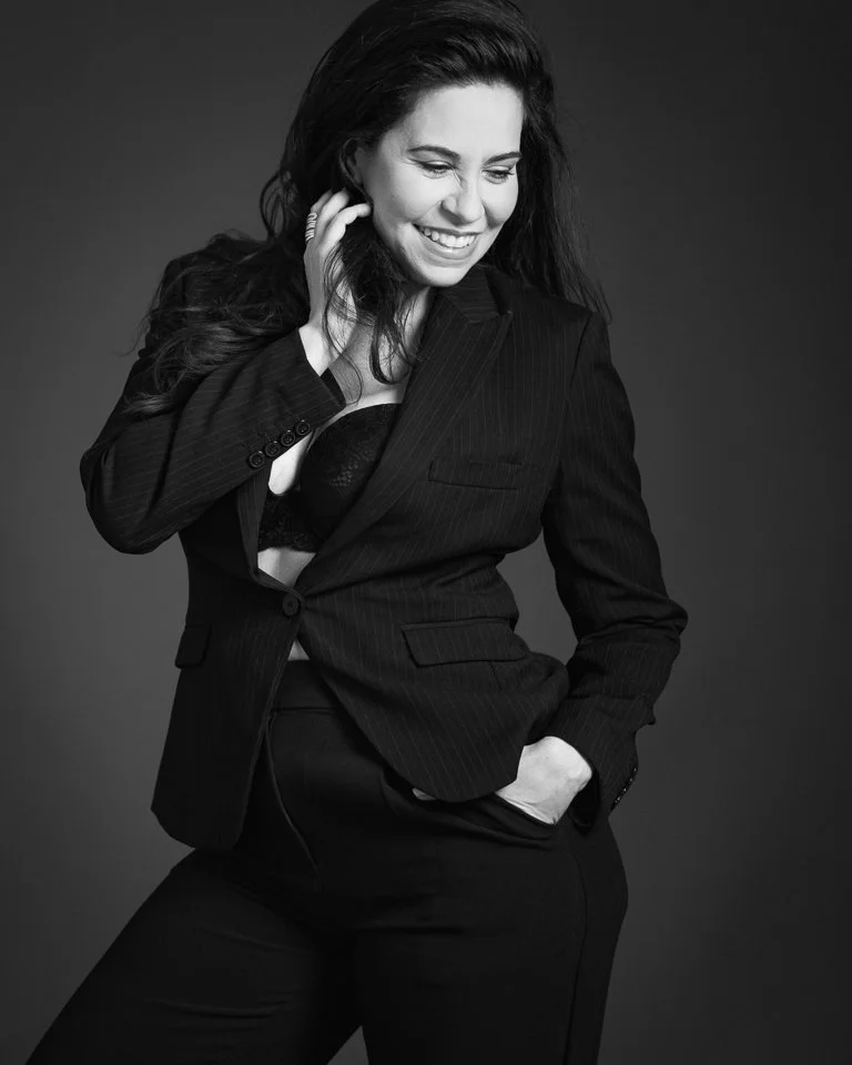 A smiling woman with dark, wavy hair wearing a pinstripe suit, lifting her jacket to reveal a lace top, with her left hand in her pocket, against a dark background.