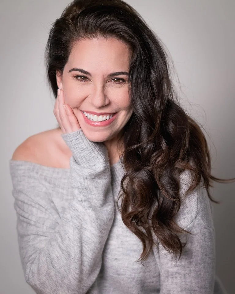 A woman with long dark wavy hair, smiling, wearing a light gray off-the-shoulder sweater, touching her face with her left hand.
