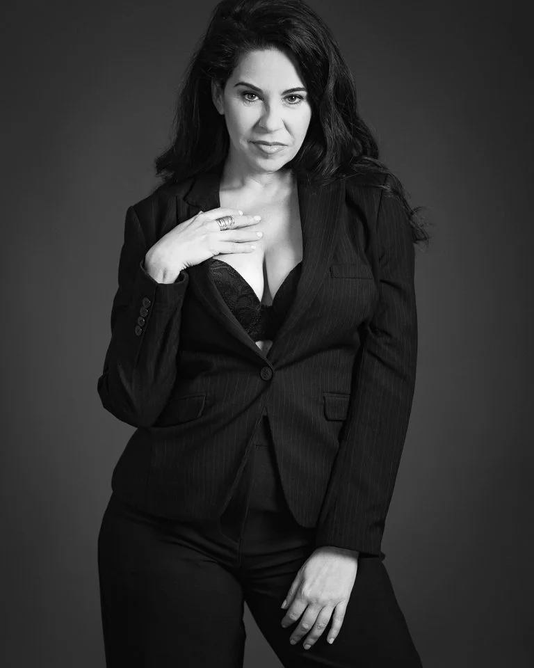 Black and white portrait of a woman with dark hair wearing a pinstripe blazer, lace top, and pants, with one hand touching her chest and the other resting on her thigh, looking at the camera.
