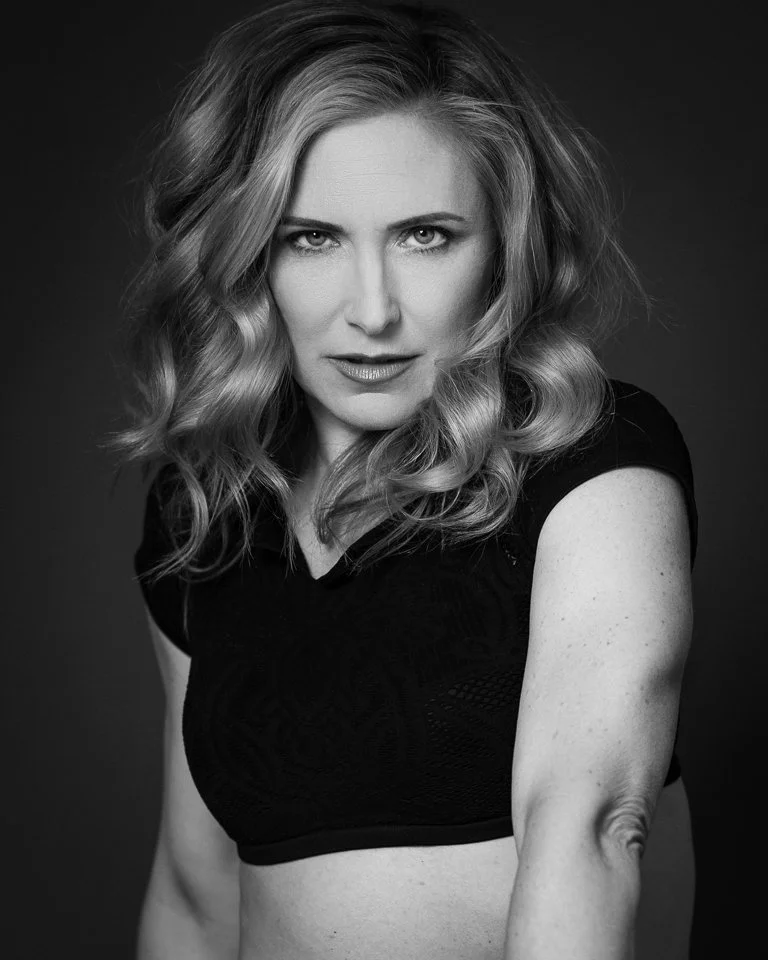 Black and white photo of a woman with wavy hair wearing a dark top, looking directly at the camera with an intense expression.