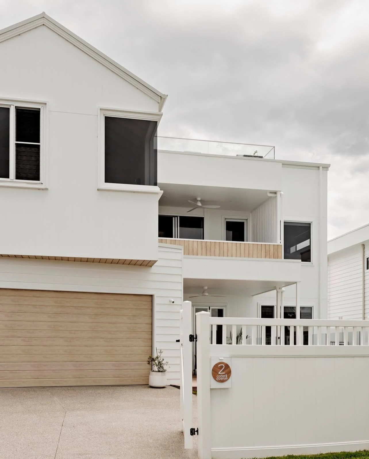Modern white two-story house with wooden garage door, front gate, and balcony, under a cloudy sky.