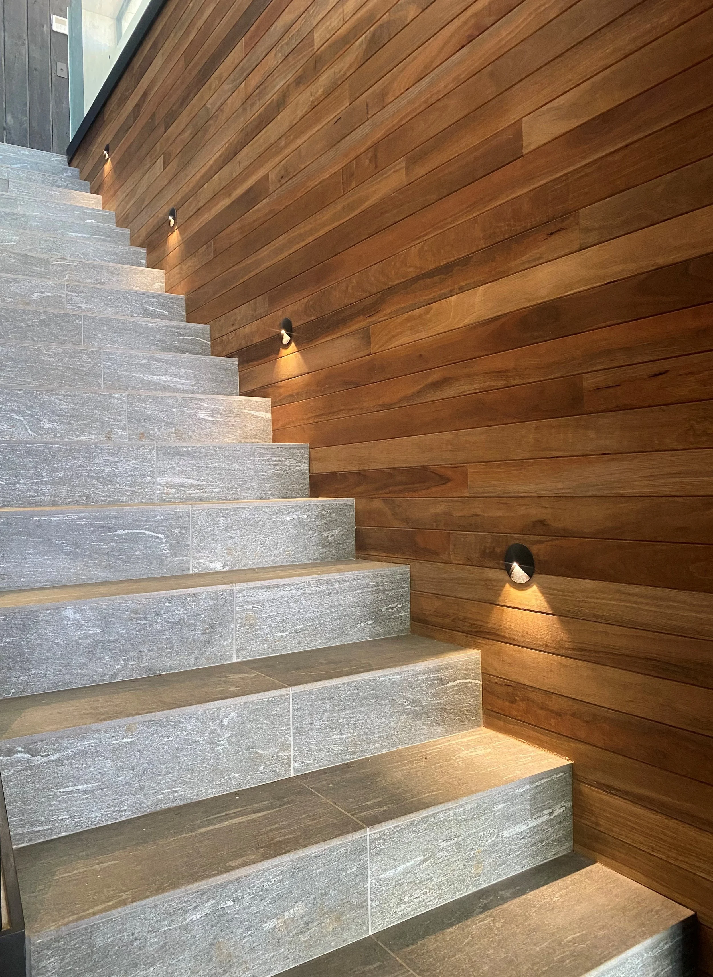 Indoor staircase with gray stone steps, illuminated by small wall-mounted lights, next to a wooden-paneled wall.