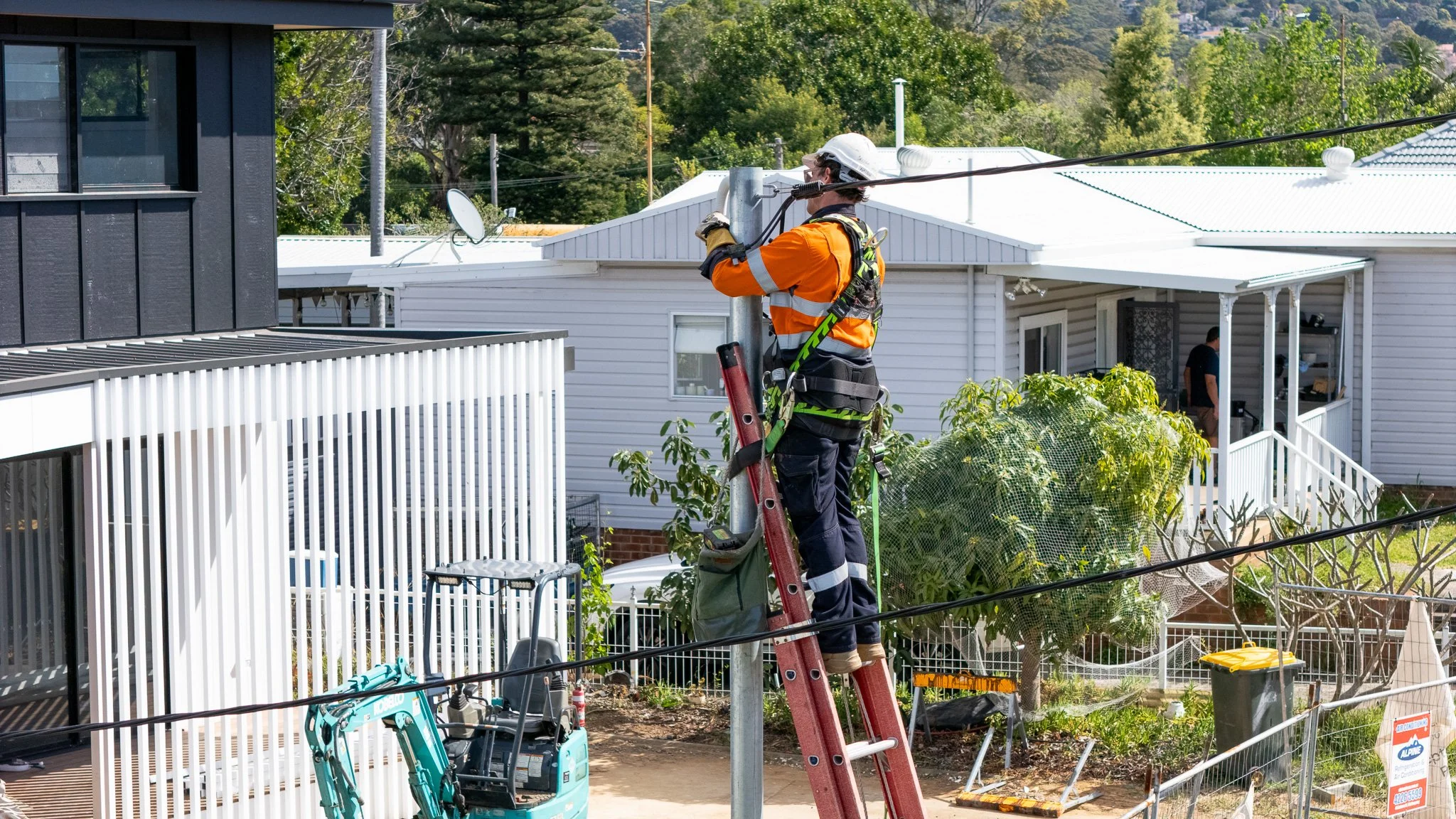 A utility worker wearing a hard hat and safety gear standing on a ladder, working on a streetlight or utility pole, with residential houses, trees, and equipment visible in the background.