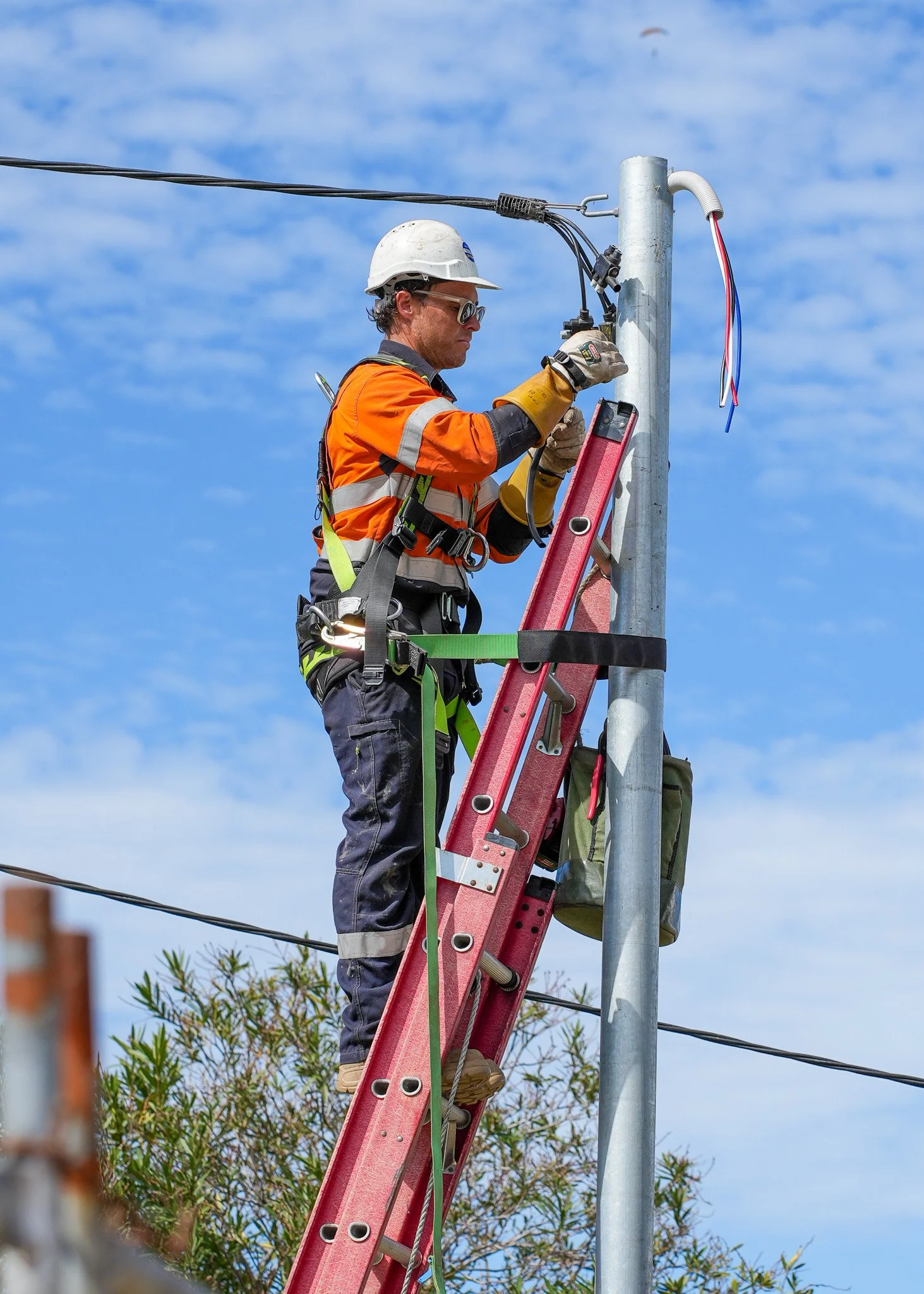 A utility worker in safety gear standing on a red ladder while working on electrical wires attached to a streetlight pole under a blue sky.
