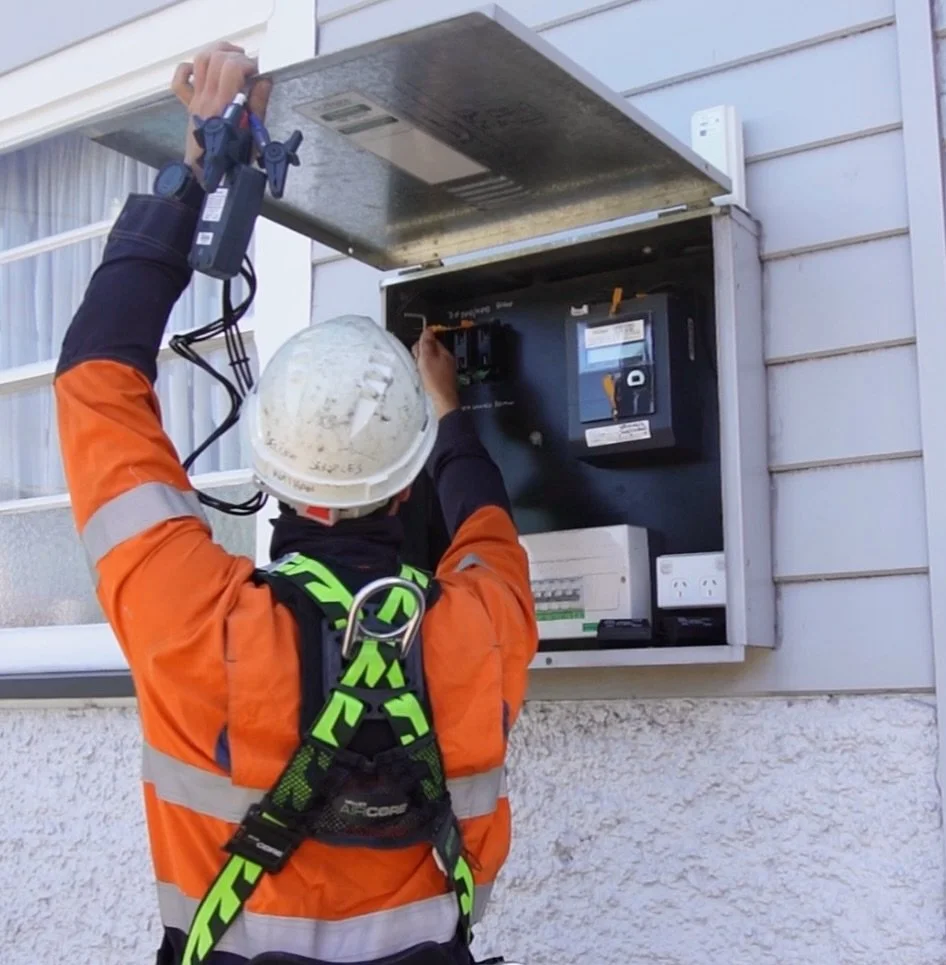 A worker wearing an orange safety jacket, a white hard hat, and a safety harness is repairing or inspecting an electrical meter box on the exterior wall of a building.