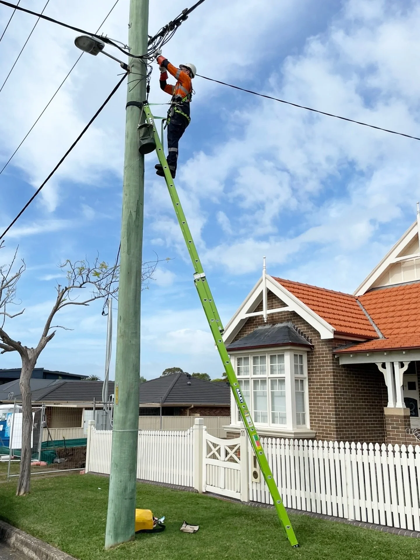 A worker on a green ladder repairing power lines on a utility pole in a residential neighborhood with houses, a white picket fence, and a tree, under a partly cloudy sky.