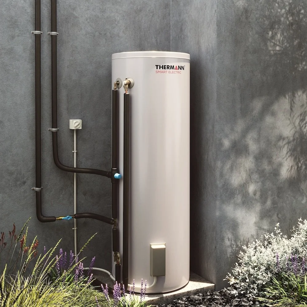 A white electric water heater with black pipes and fittings installed outdoors against a concrete wall, surrounded by plants and shrubs.