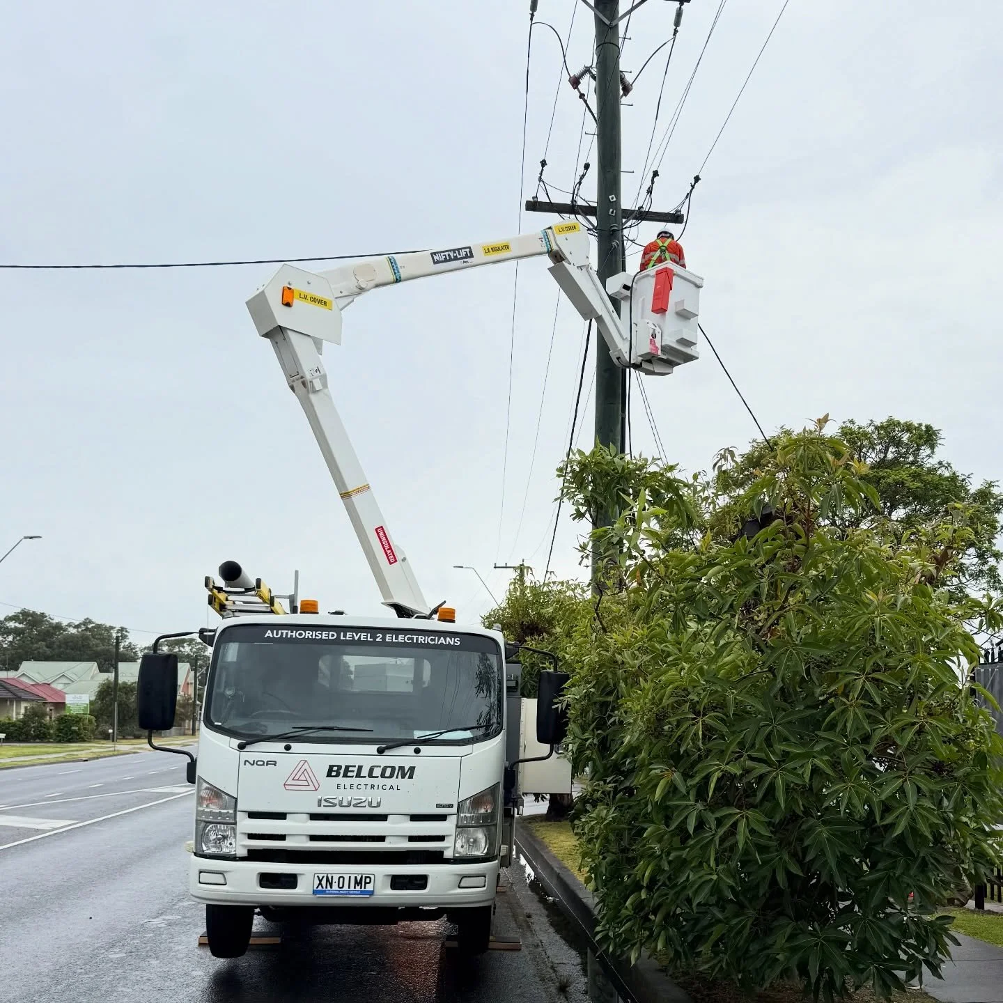 A utility worker in an orange safety vest in a bucket lift inspecting power lines on a utility pole. A white service truck with the label 'BELCOM Electrical' and 'Authorised Level 2 Electricians' is parked beside a sidewalk.