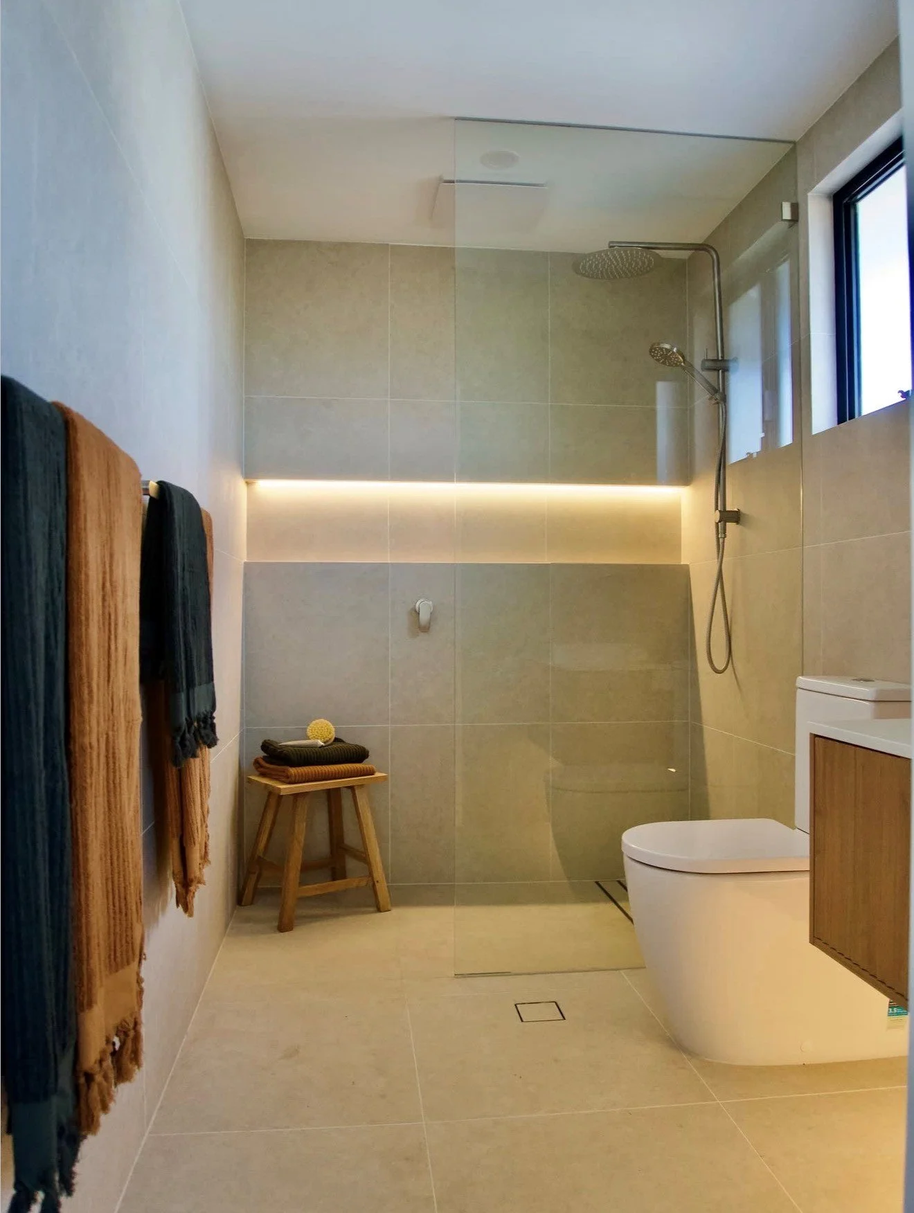 Modern bathroom with beige tiles, a walk-in shower with a rain showerhead, a small window, towels hanging on a rack, a small wooden stool with folded towels, and a white toilet with a wooden cabinet.