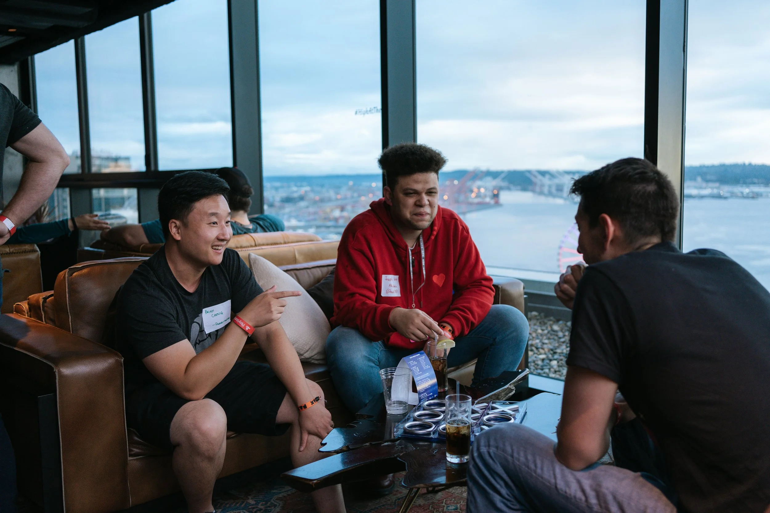 Three men sit talking together on couches in hotel event space, large windows in background show Seattle skyline