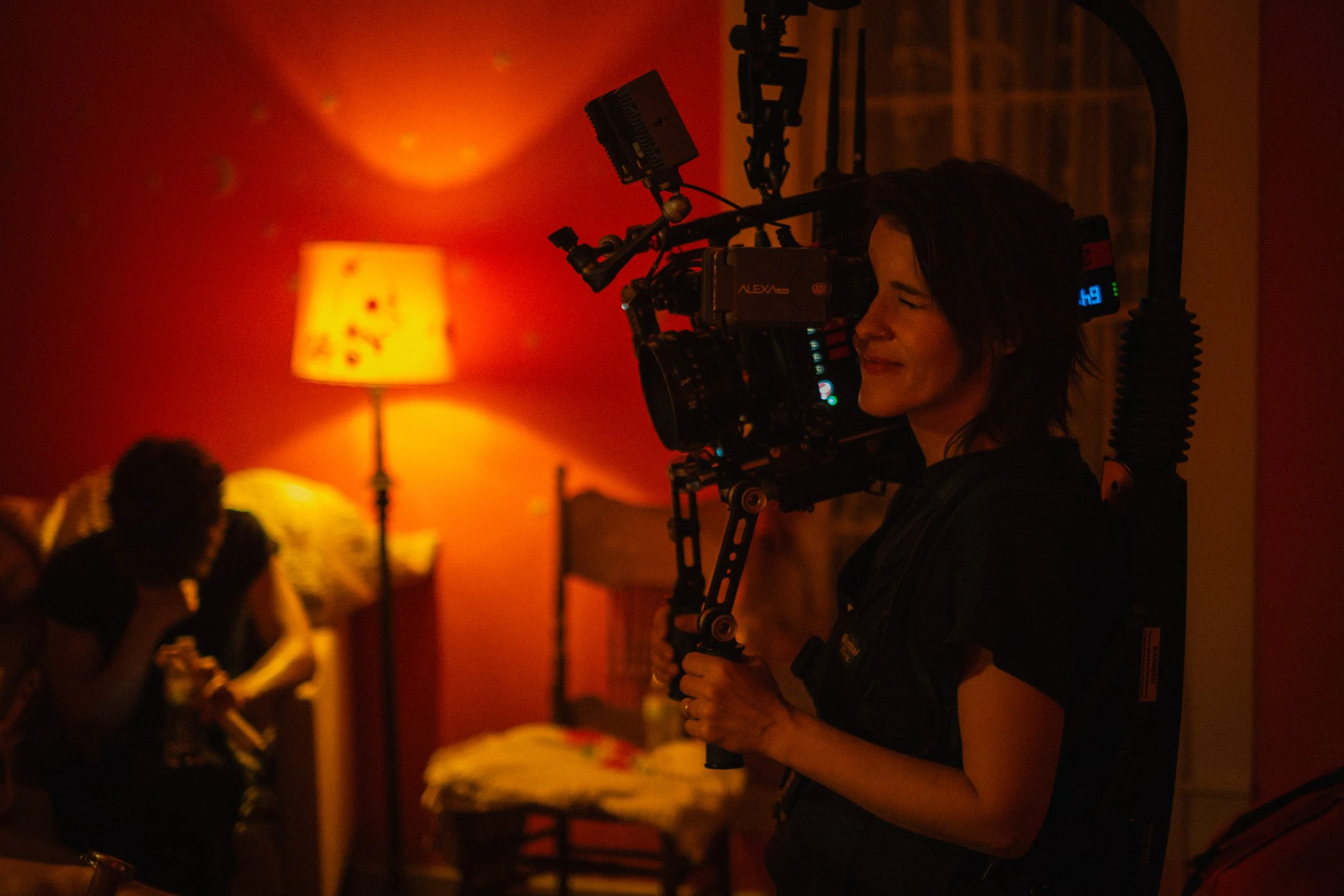 In dim, warm, light, female cinematographer Ashton McKenzie operates ARRI Alexa Mini on an EasyRig on set of independent feature film Openers
