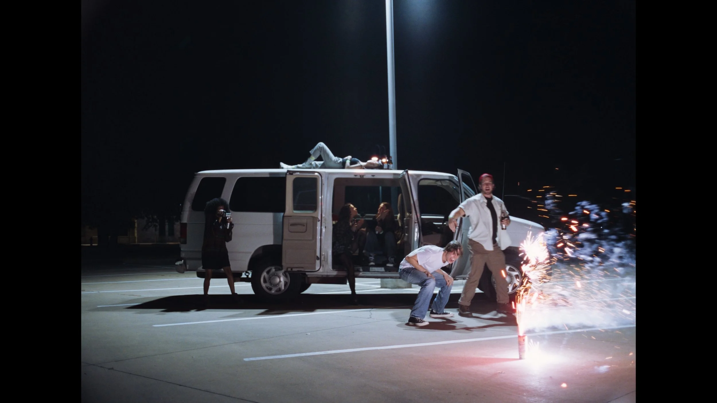 cinematic frame grab shows group of young people hang outside van parked under single street light in empty parking lot, watching ground firework blooming