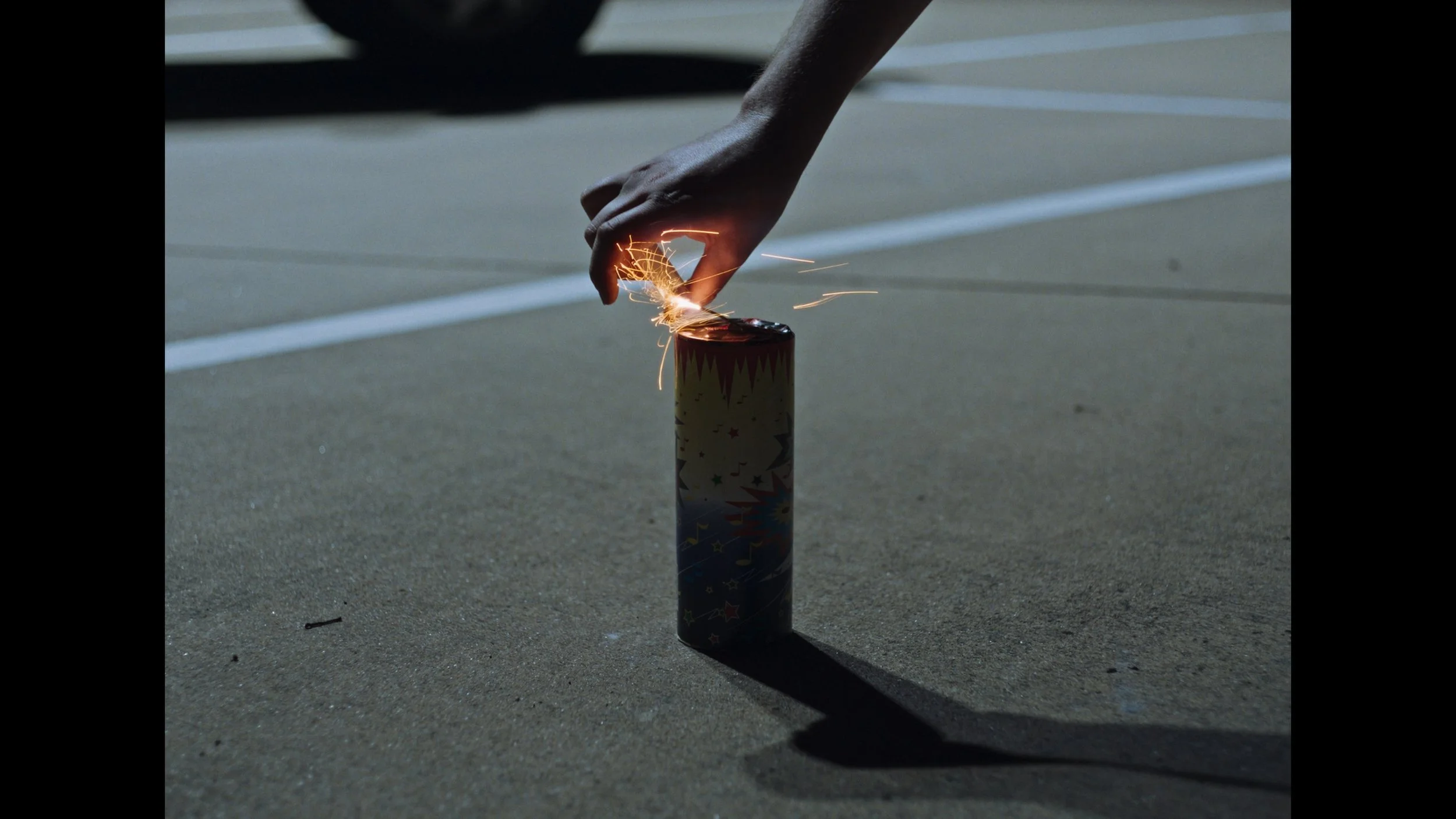 frame grab from the indie film Openers, a firework sits on the ground of a parking lot, a hand reaches down to light it