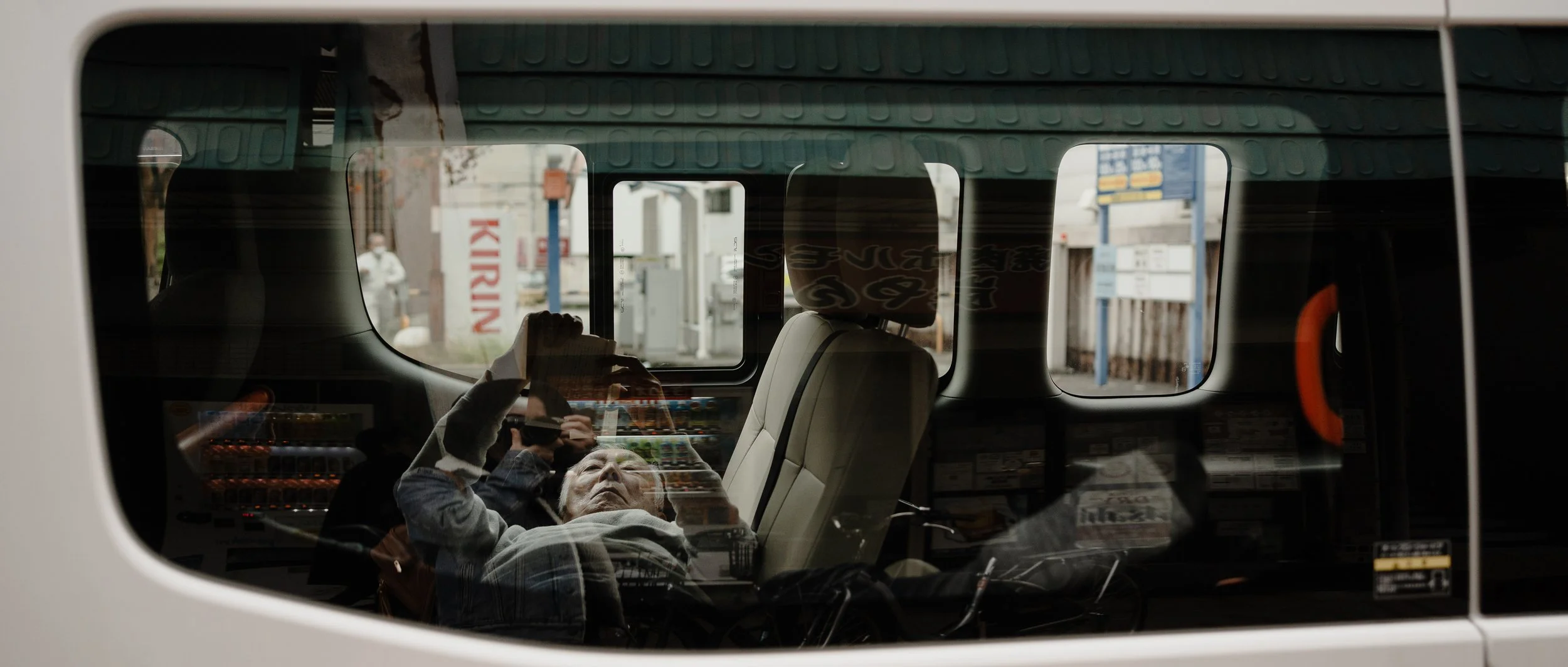 Looking in through window, man lays down across seat in van, holds phone overhead to read, Kirin vending machine in background situates the scene in Japan