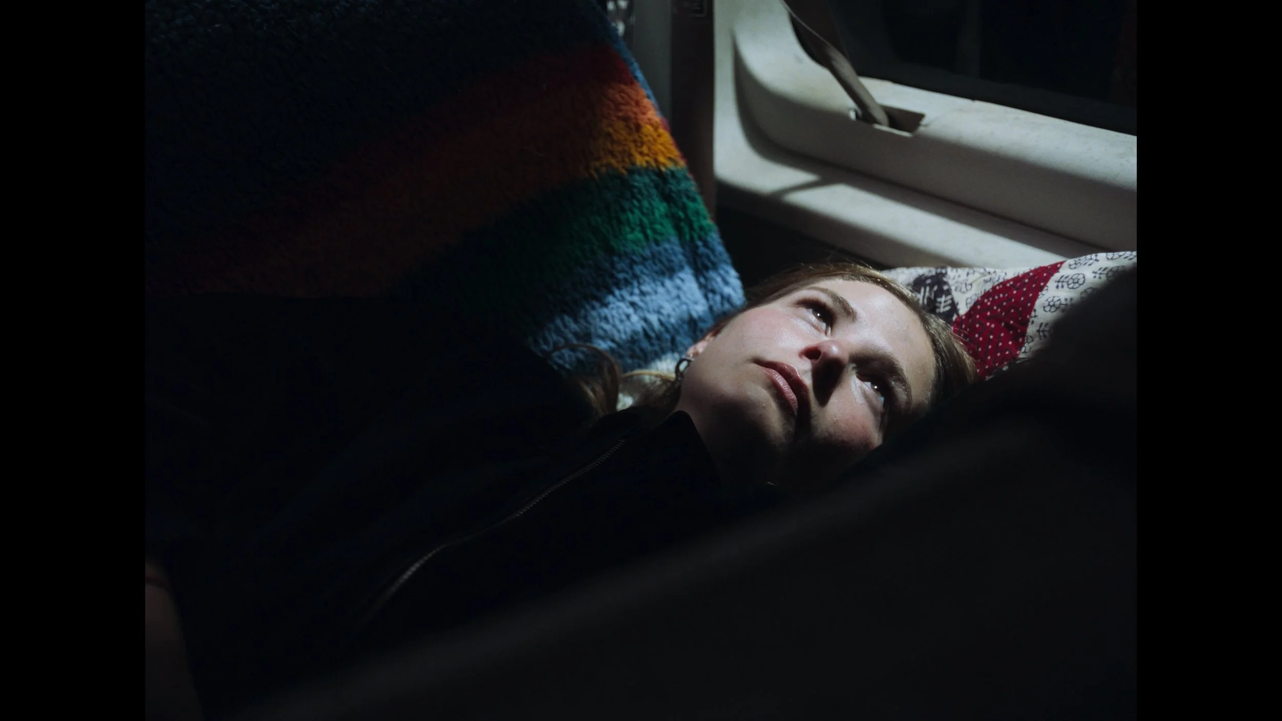 cinematic still frame from indie film, Openers, woman lays on bench seat in back of a van at night, street lights illuminate her face as she looks out the window