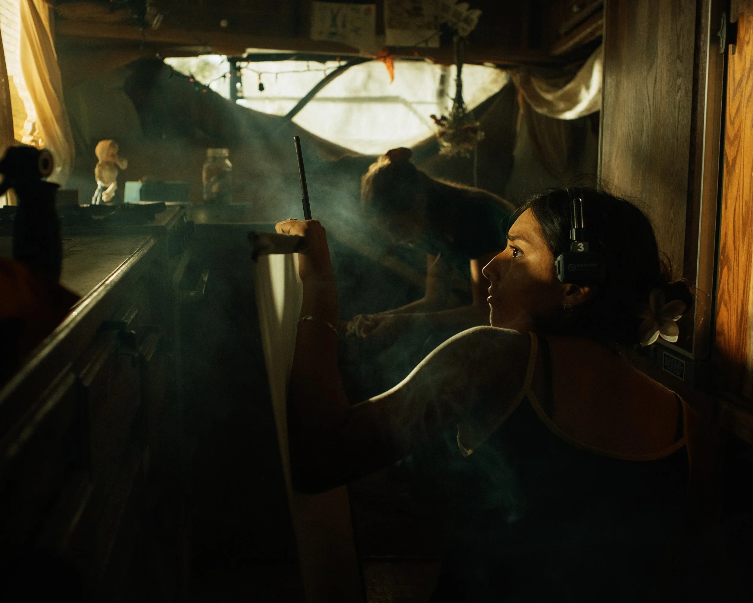 Female grip on film set looks into the light, fans away artificial haze, actress in background prepares to begin filming