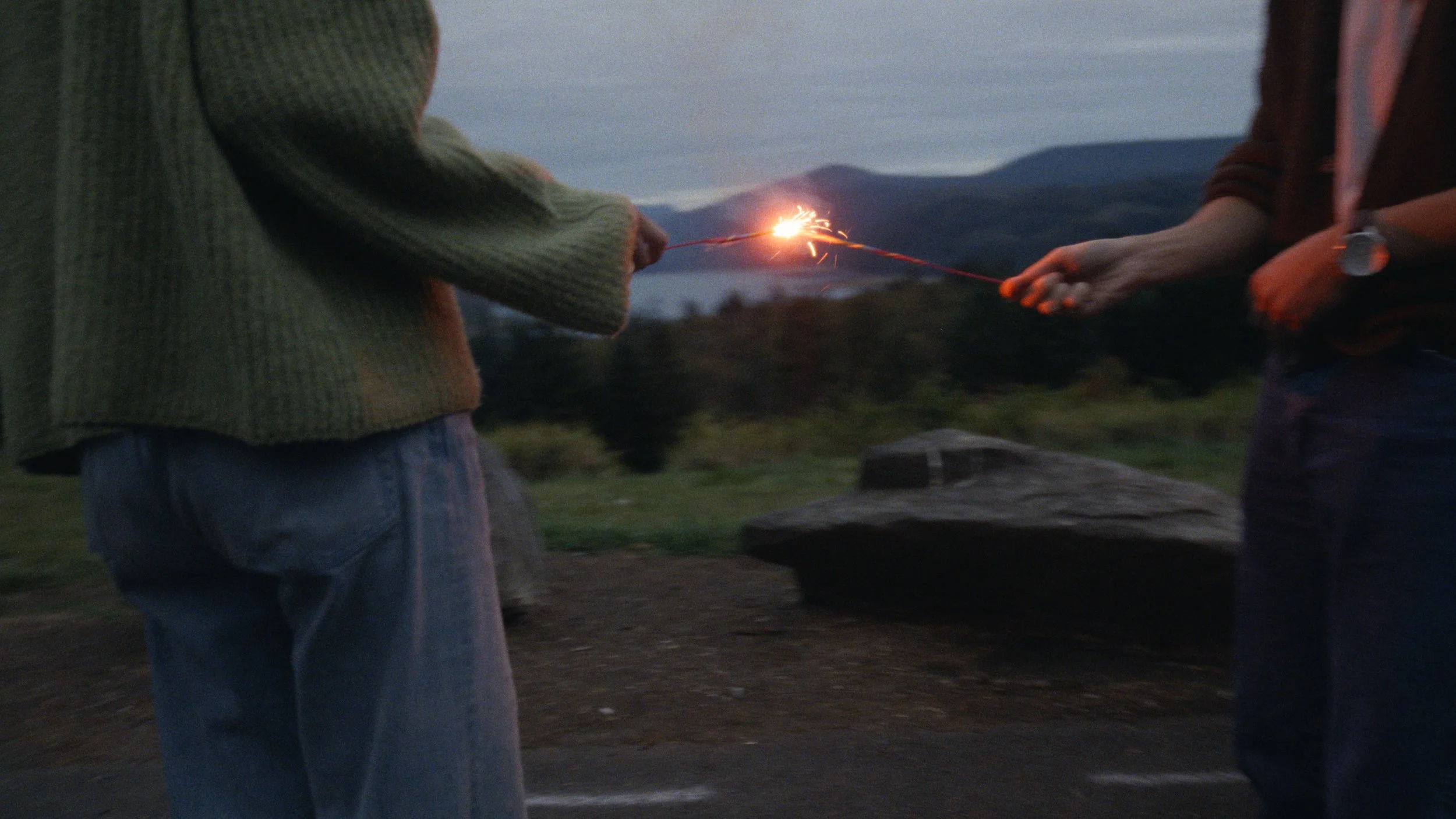 Cinematic frame grab, medium close up of hands holding sparklers as they get lighted, backdropped by a lake and mountain range at blue hour