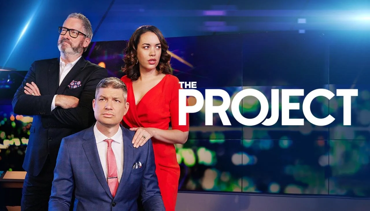 Promotional image for "The Project" featuring three people: a bearded man in glasses with arms crossed, a woman in a red dress looking left, and a clean-shaven man in a suit seated with a serious expression. "The Project" text is on the right.