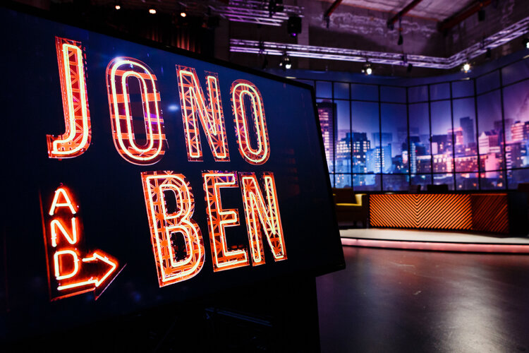Television studio set with a large illuminated sign reading "Jono and Ben" in neon-style letters. The background shows a nighttime cityscape backdrop with a talk show desk and seating area.