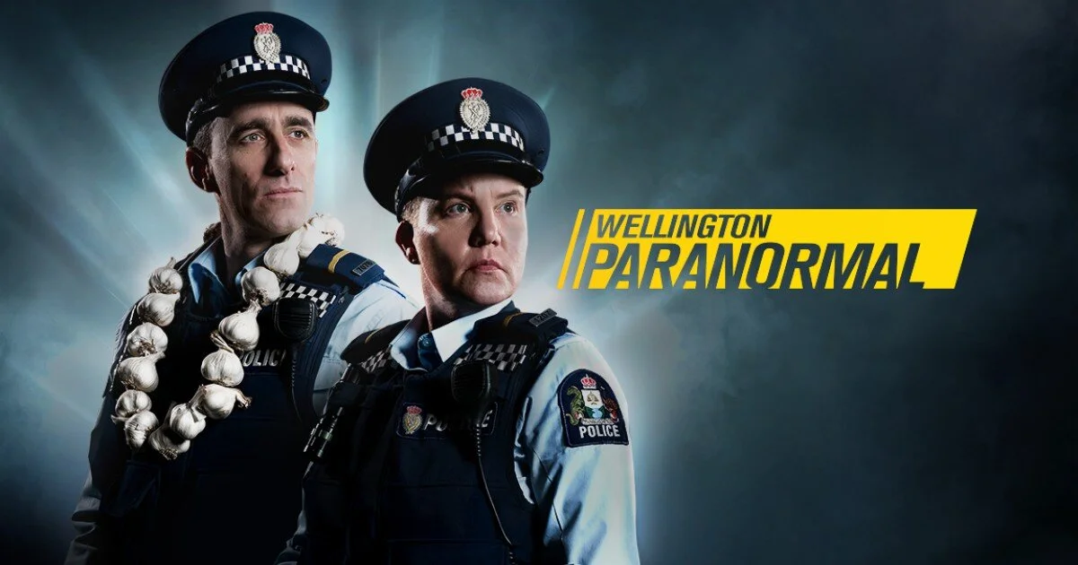 Promotional poster for TV show "Wellington Paranormal" featuring two police officers, one wearing a garlic necklace, with the show's title in yellow text.