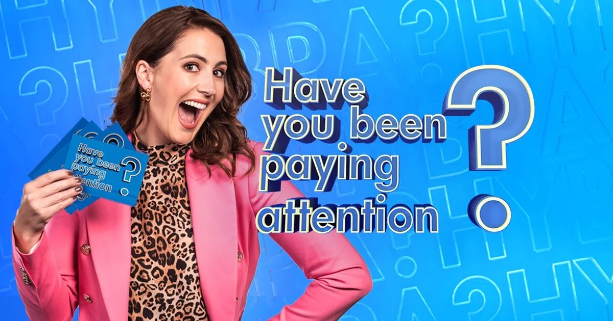 Excited woman in pink blazer holding cards with text "Have you been paying attention?" on blue background.