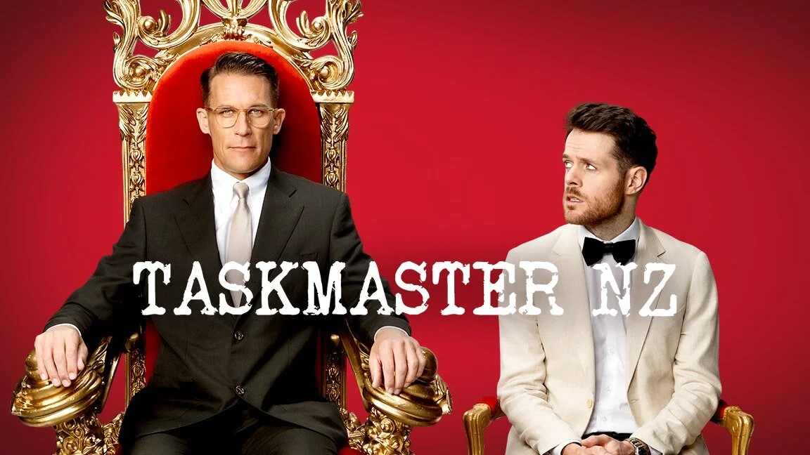 Promotional image for "Taskmaster NZ" featuring two men in formal attire; one seated on a throne, the other standing beside him, against a red background.